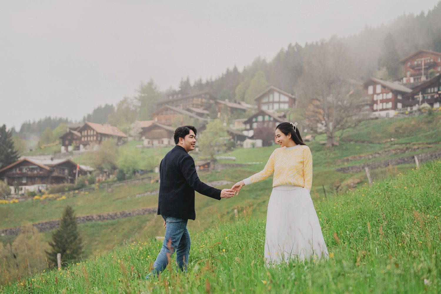 Couple holding hands and walking through a lush green meadow with misty Swiss village behind.