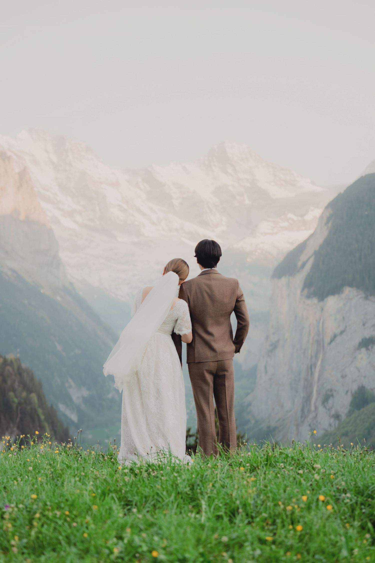 Bride leaning on groom's shoulder as they stand on alpine meadow overlooking majestic Swiss mountain valley.