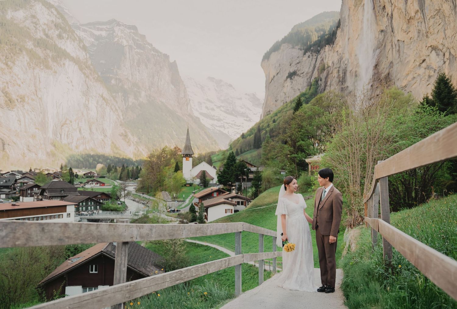 Bride and groom standing on a wooden bridge overlooking the picturesque village of Lauterbrunnen, Switzerland.