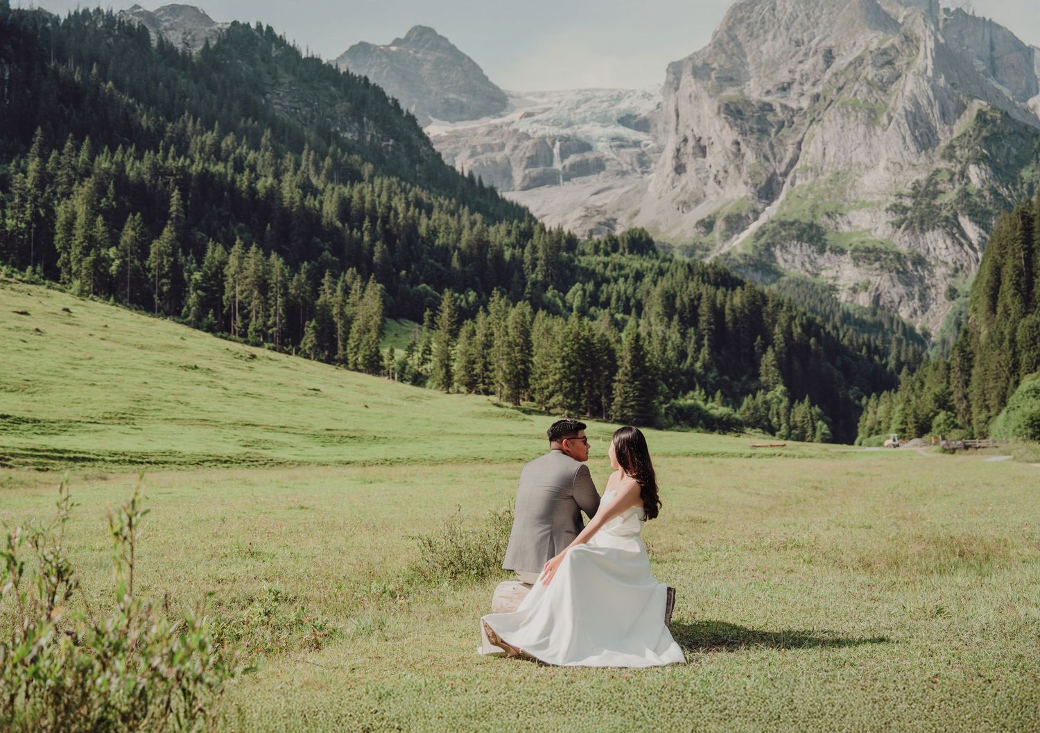 Bride and groom sitting together in lush green alpine meadow with mountain glacier background.
