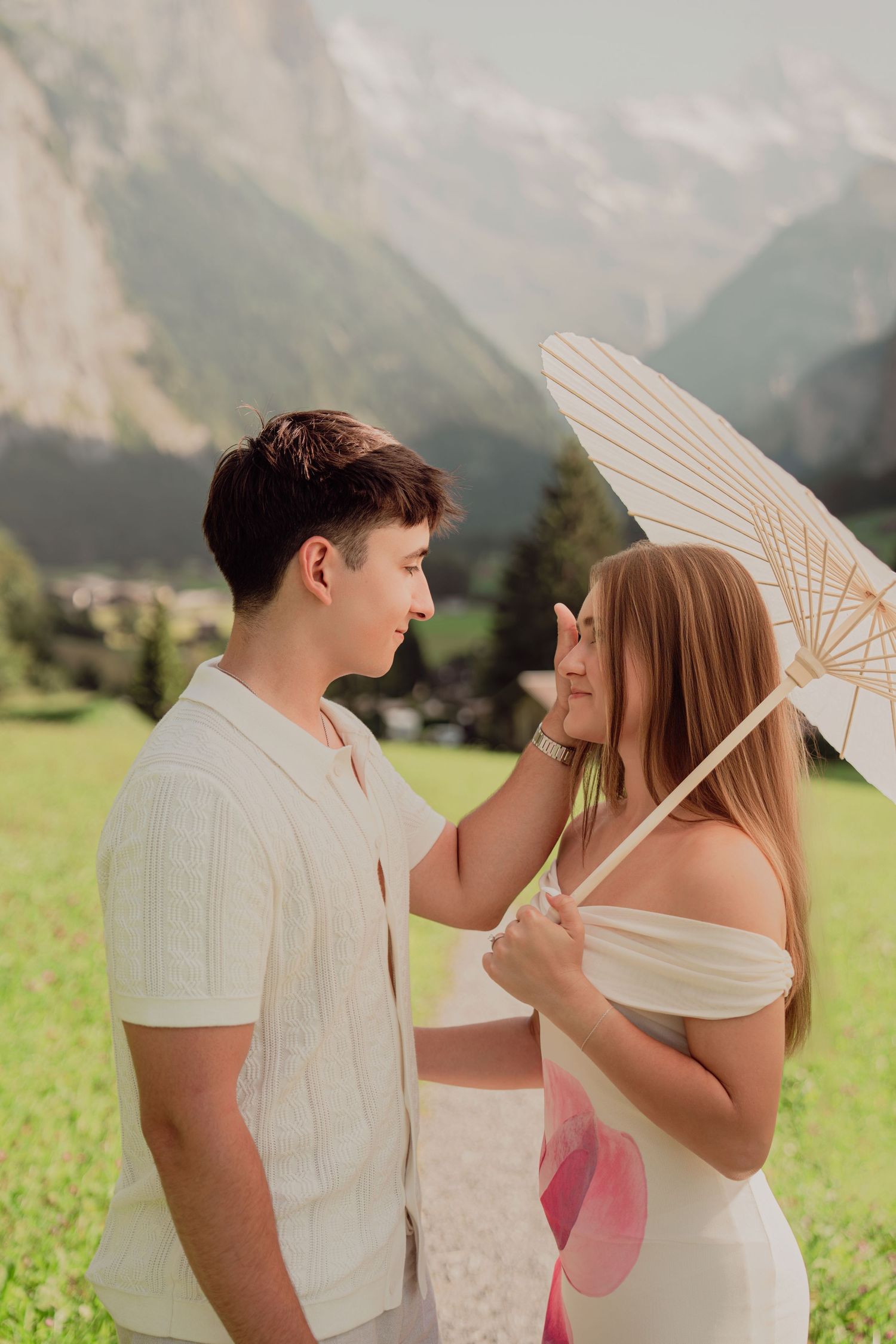 Young couple smiling at each other under elegant white paper umbrella with stunning alpine mountain scenery behind them.