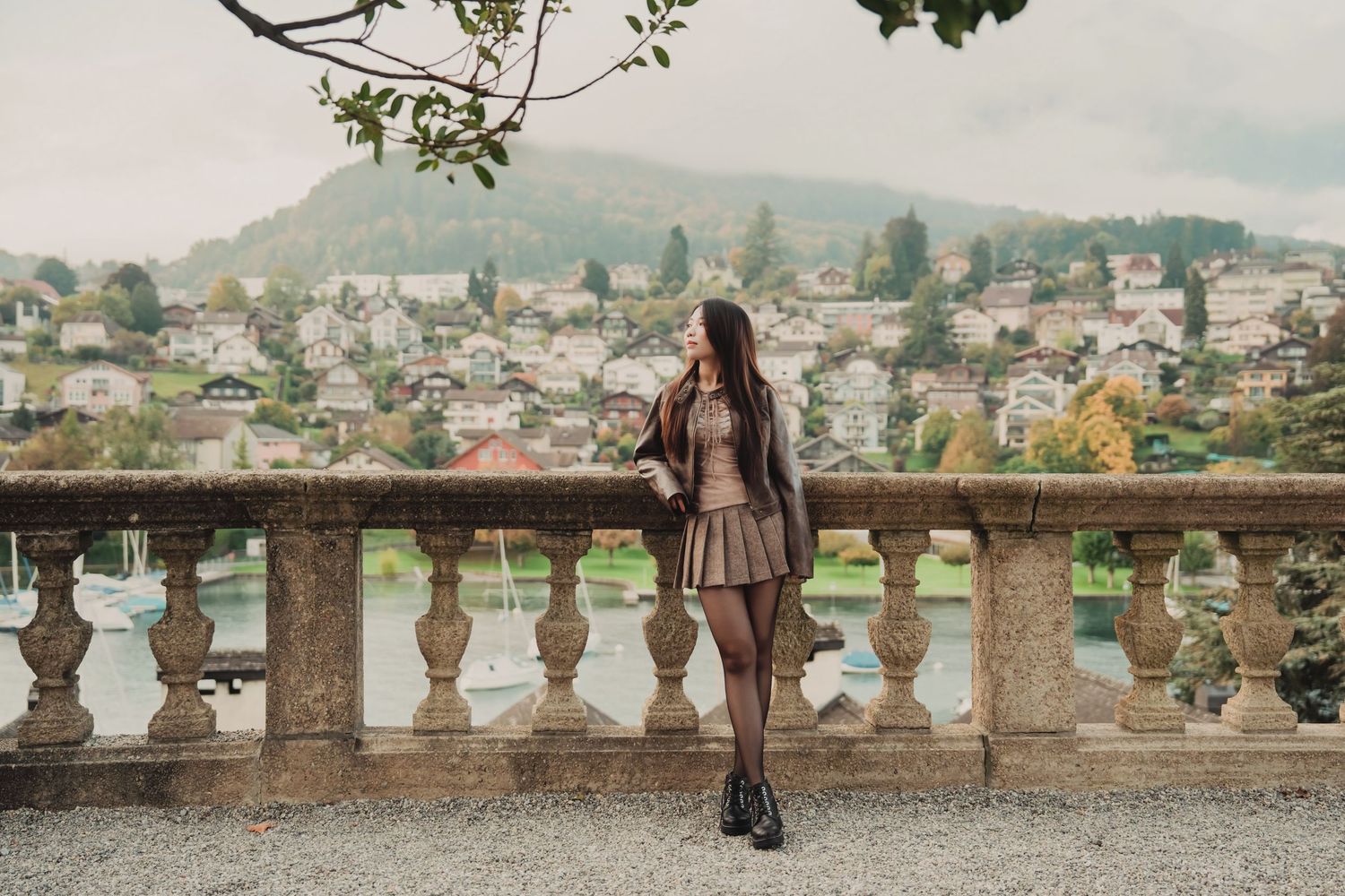 Elegant woman in a brown jacket and skirt leaning on a stone balustrade with a scenic town view behind.