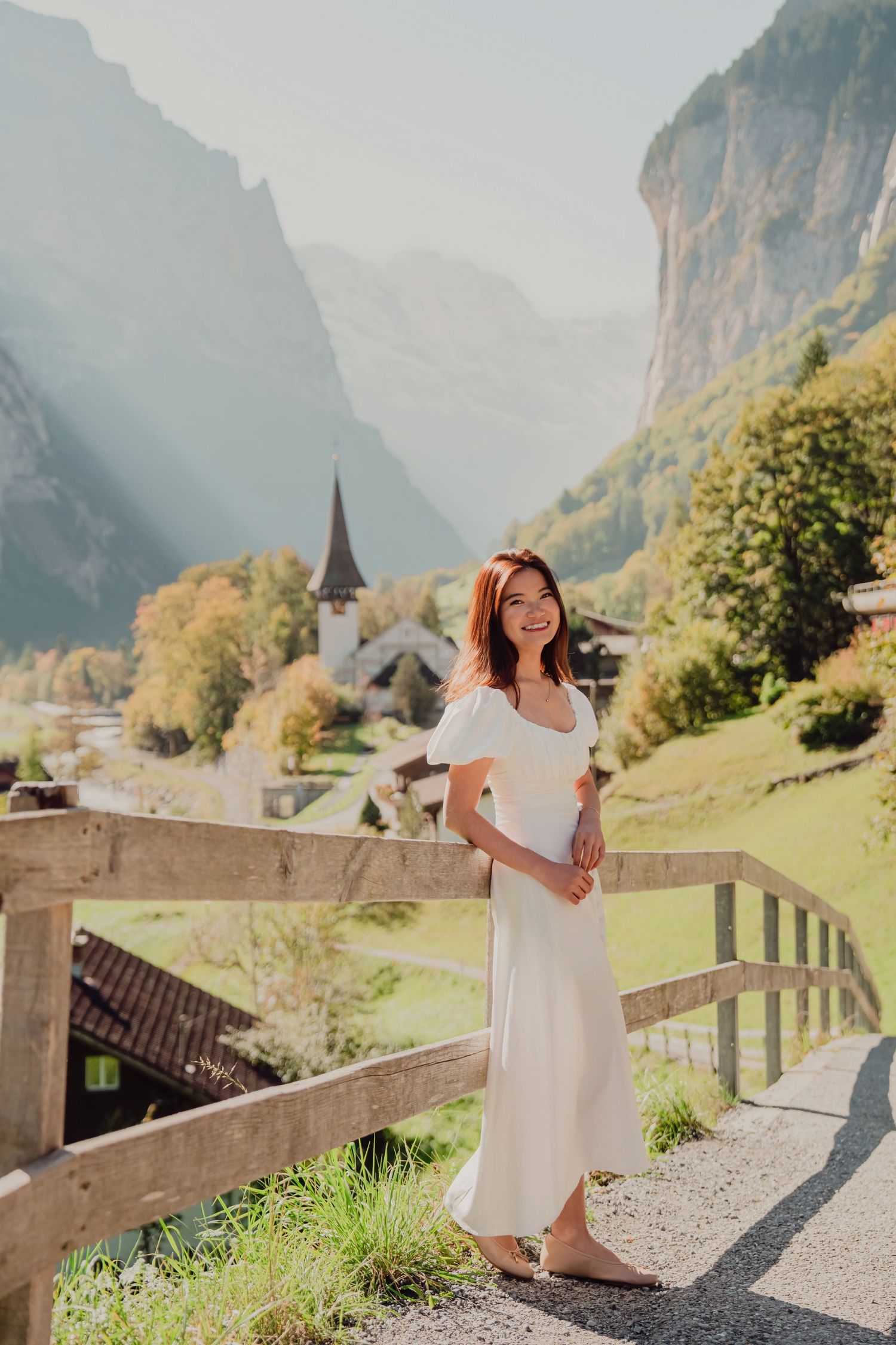 A happy woman in a white dress leans on a wooden fence with a scenic Swiss mountain village behind her.