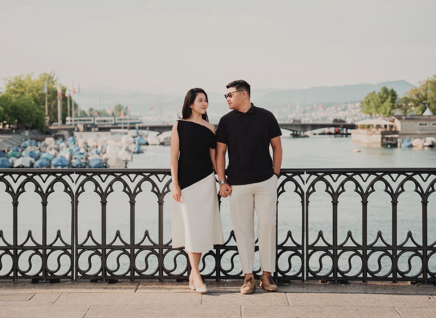 Couple holding hands by ornate iron railing with Zurich lake and cityscape in background.