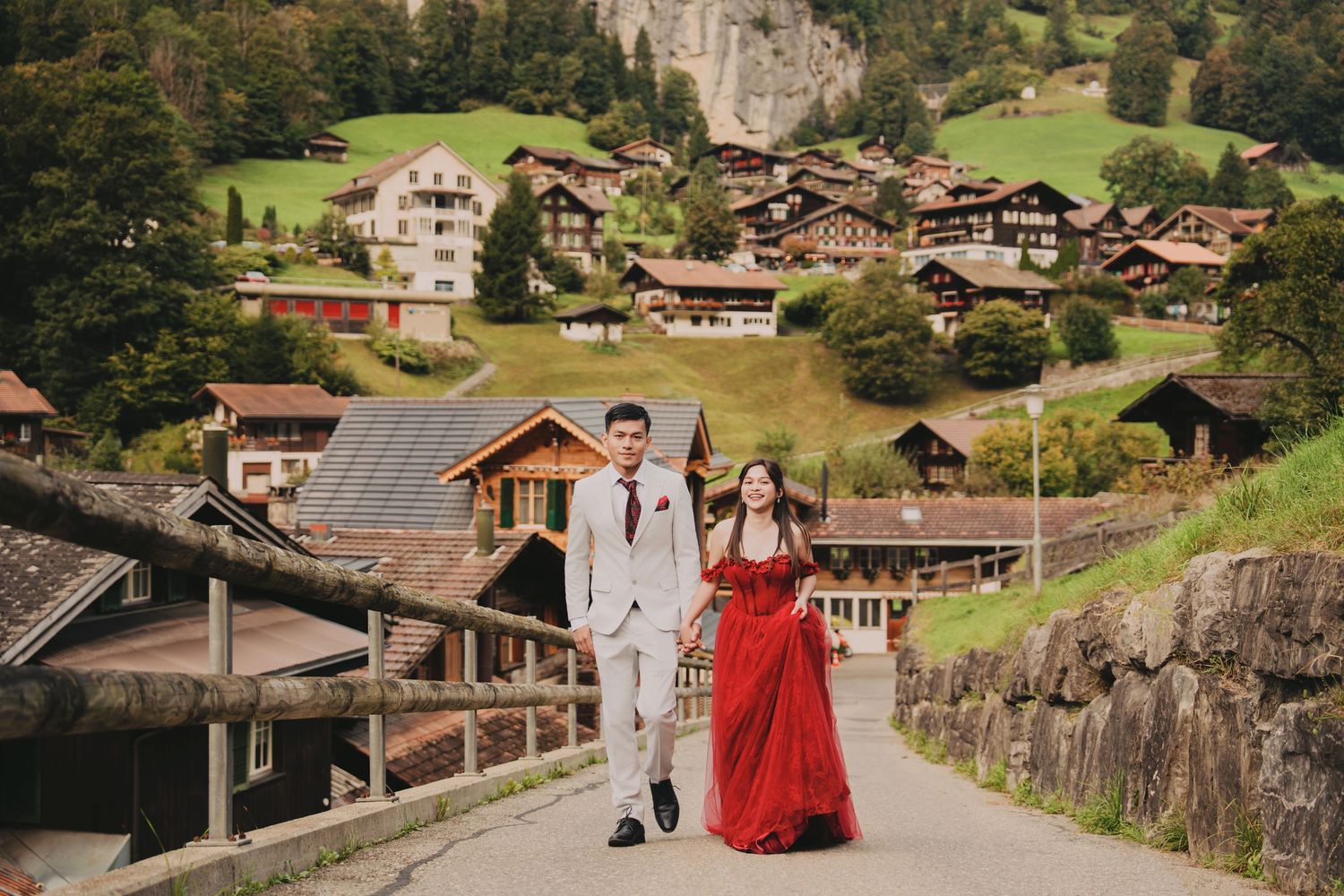 Elegant couple strolling through a scenic Swiss mountain village, bride in red off-shoulder gown.