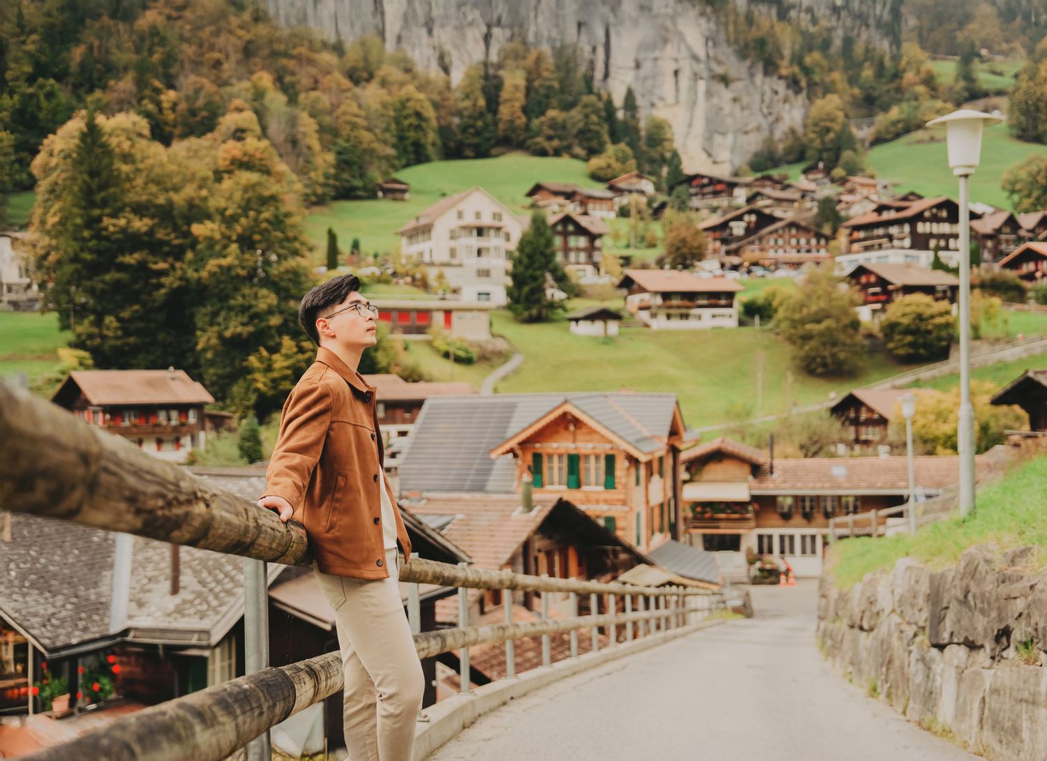 Young man in brown jacket standing on a bridge overlooking a scenic Swiss alpine village road in autumn.