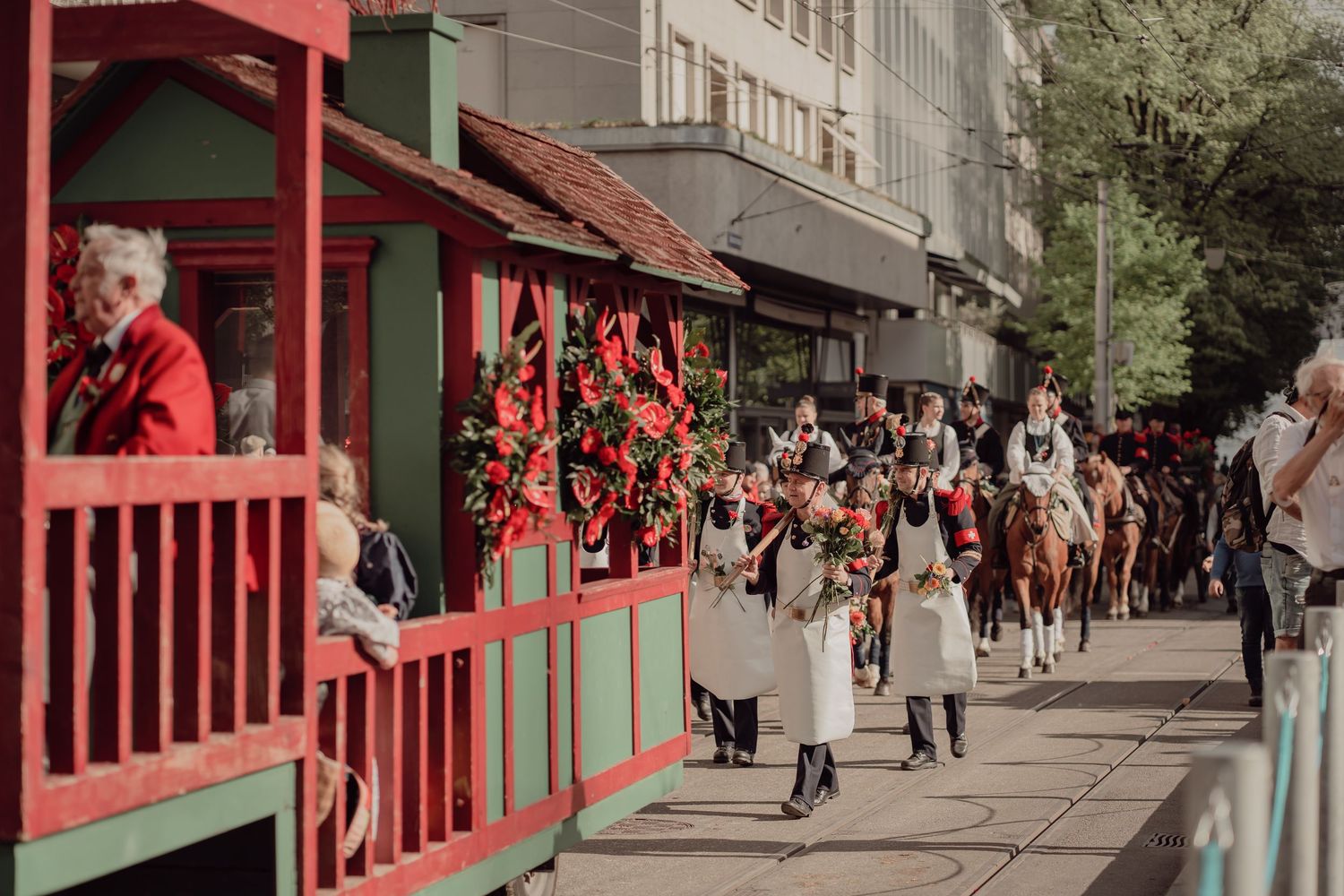 Historical parade procession with horse-drawn red tram decorated with flowers moving through city street.