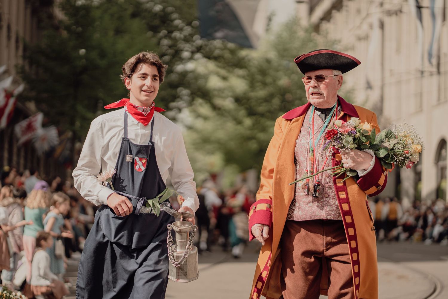 Two men in historical costumes walk in a parade, one holding a bouquet of flowers.