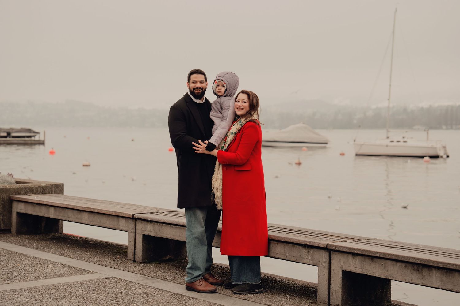 Family of three poses by a misty lakeside, parents in black coat and red coat holding their toddler.