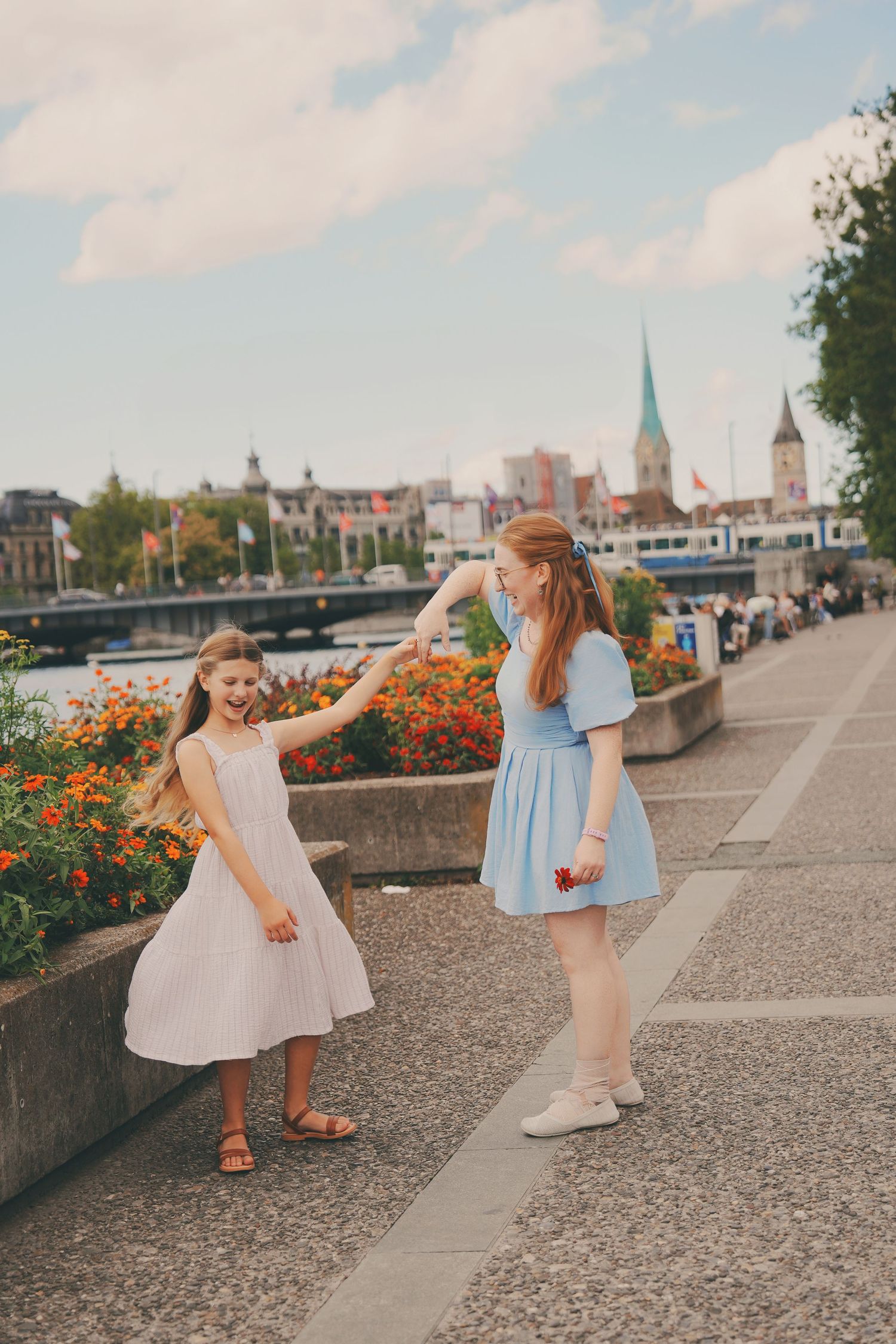 Two girls in dresses dancing and holding hands by colorful flowers along the Zurich promenade.