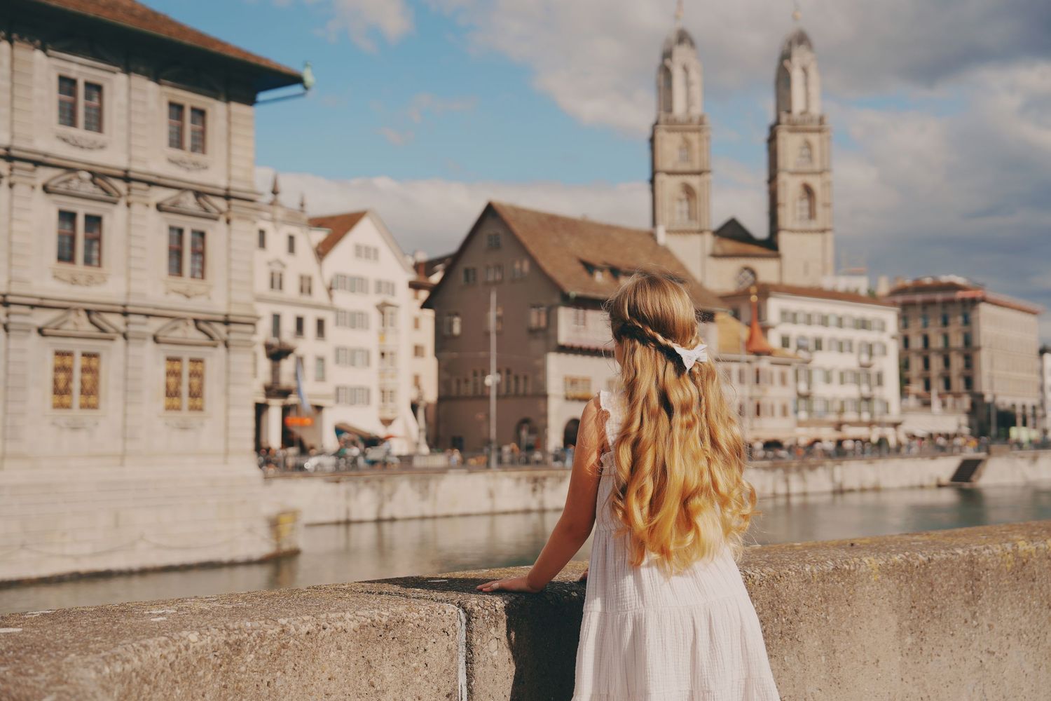 Blonde girl in white dress gazes at Zurich cityscape with Grossmunster cathedral towers.