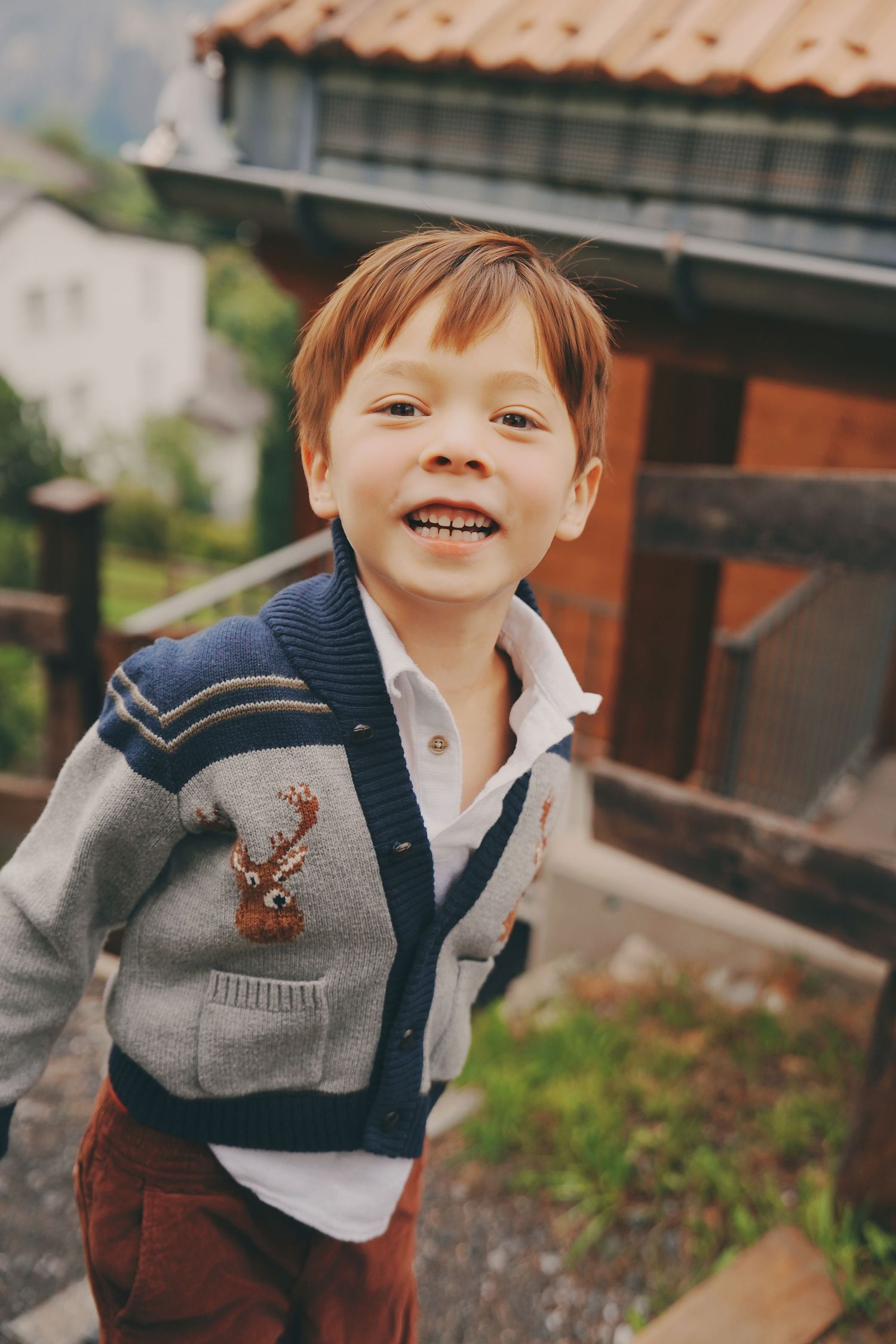 Smiling red-haired boy in a reindeer cardigan poses outdoors near alpine village buildings.