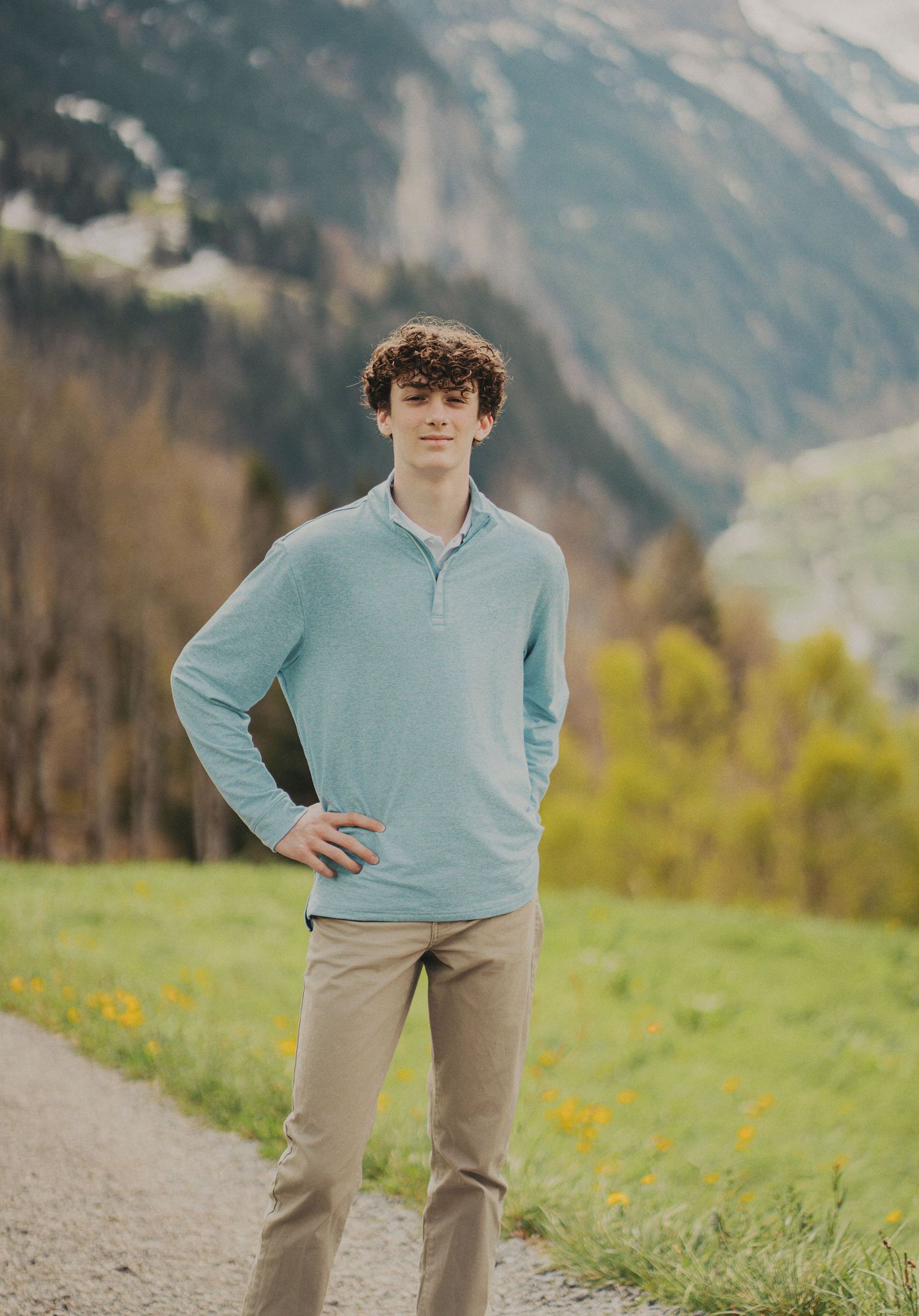 Teenage boy in light blue pullover posing confidently on a mountain path with Alpine valley scenery behind him.
