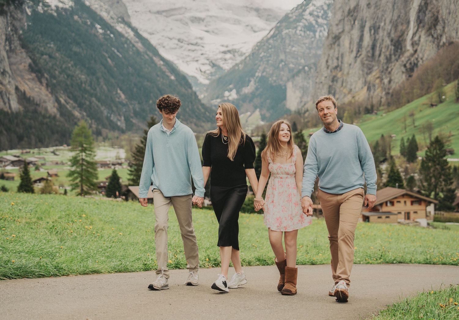 Family of four smiling and walking together on a path with snow-capped Swiss Alps in background.