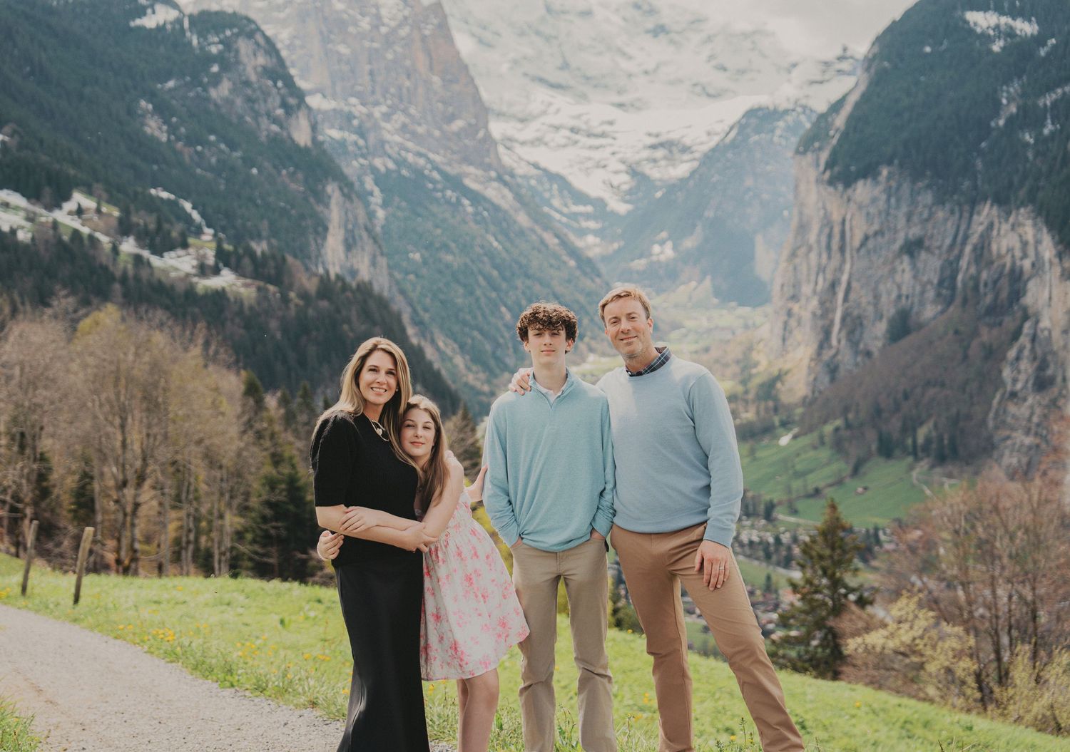 Family of four smiling together on a grassy Swiss hillside with snow-capped mountain peaks.