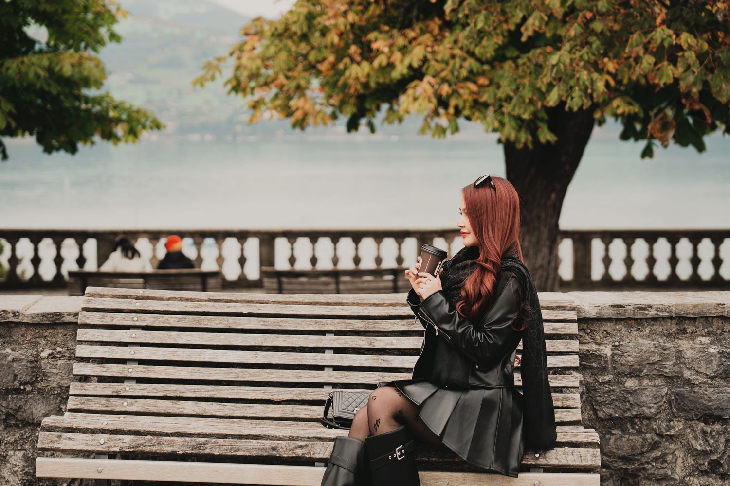 Red-haired woman in black leather jacket sits on wooden bench using smartphone near scenic autumn lakeside promenade.