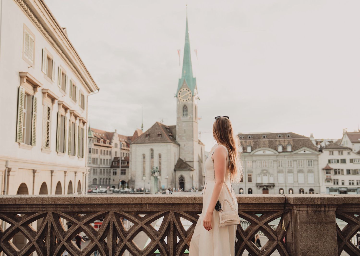 Woman in white dress admires Fraumunster Church from ornate stone bridge in old town Zurich, Switzerland.