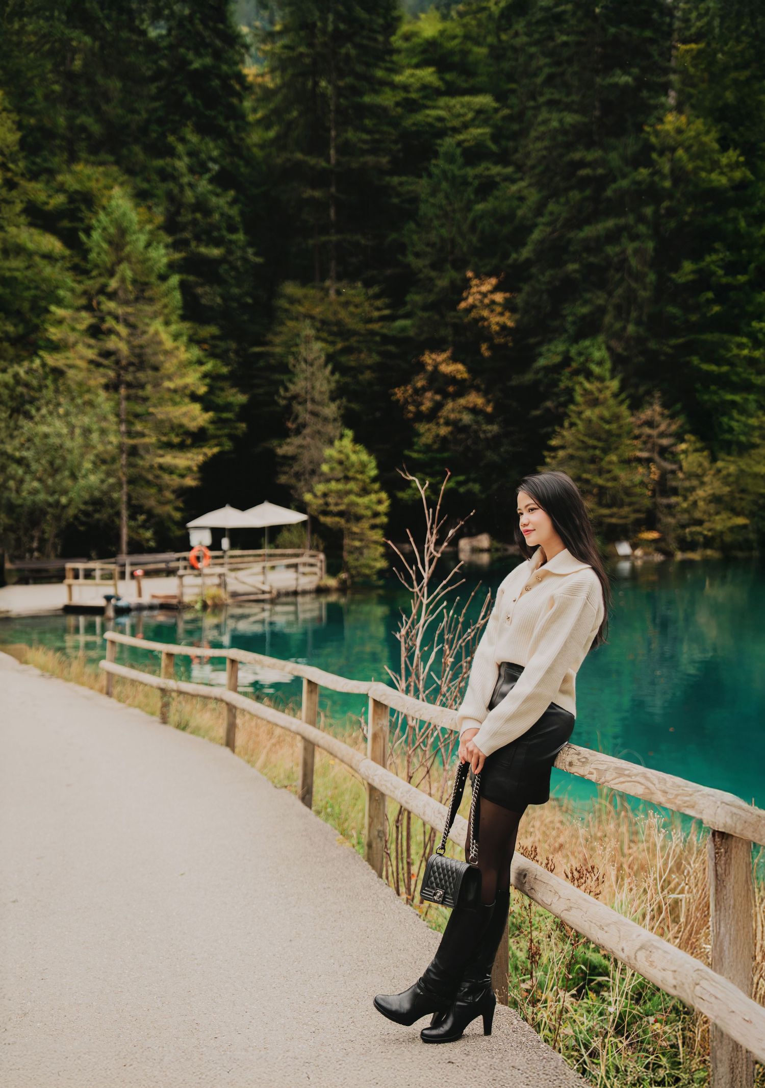 Stylish woman in white sweater and black skirt leans on wooden fence by turquoise lake.