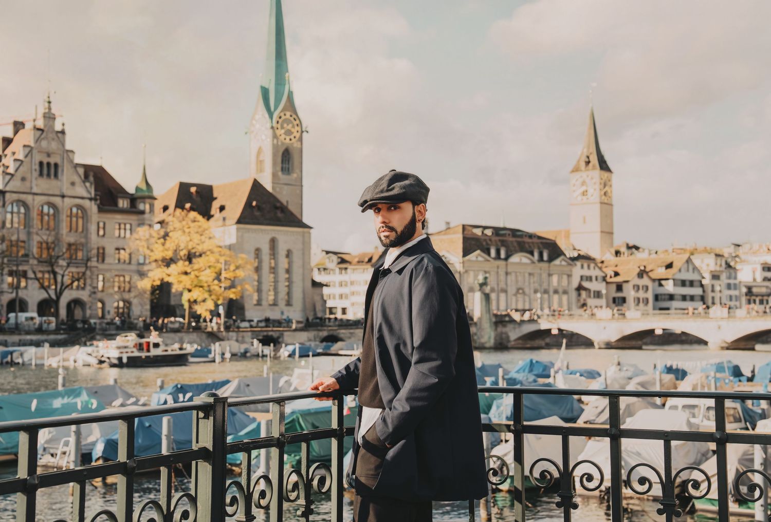 Bearded man in navy coat and flat cap stands by iron fence overlooking Zurich river and spires.