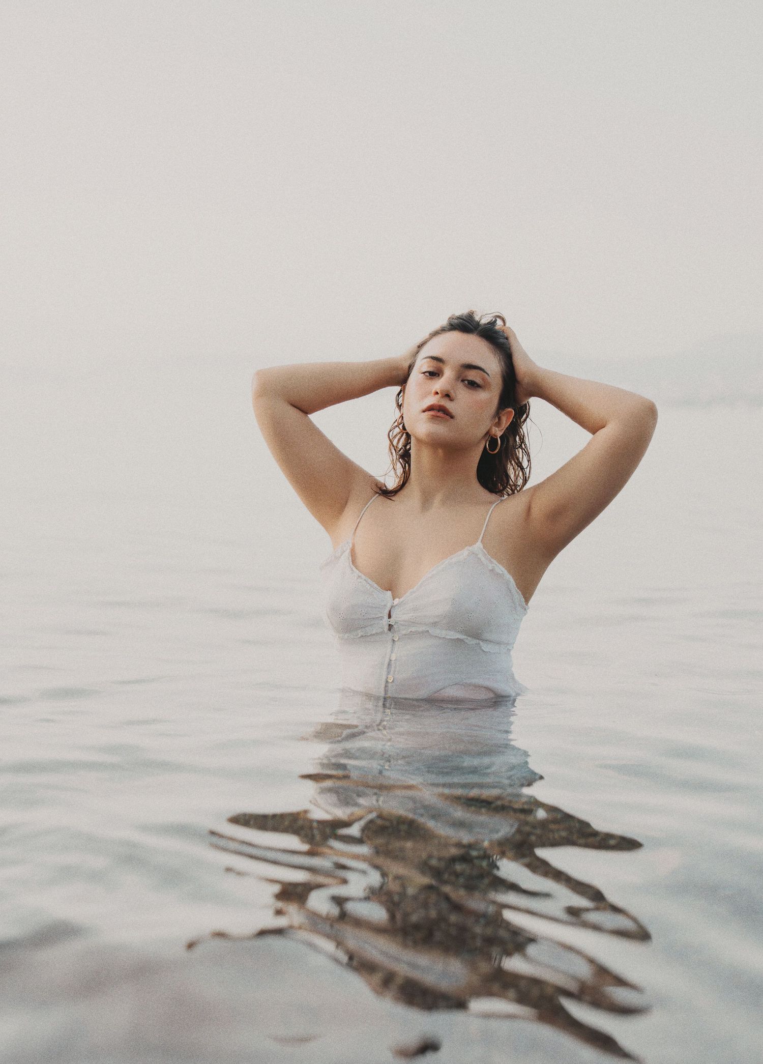 Woman in white wet camisole standing in calm ocean water with hands in hair.