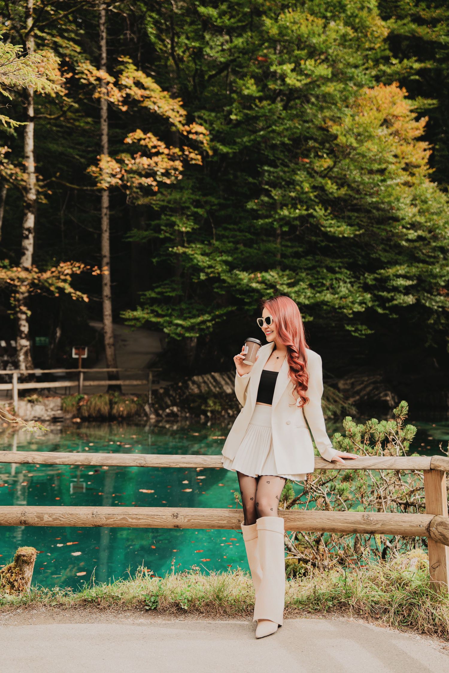 Redhead woman in white blazer holding coffee cup by turquoise lake in autumn forest.