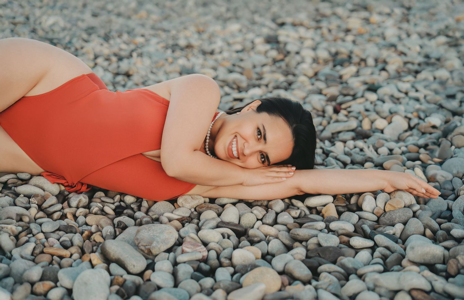 Woman in red swimsuit smiling while lying on rocky pebble beach.