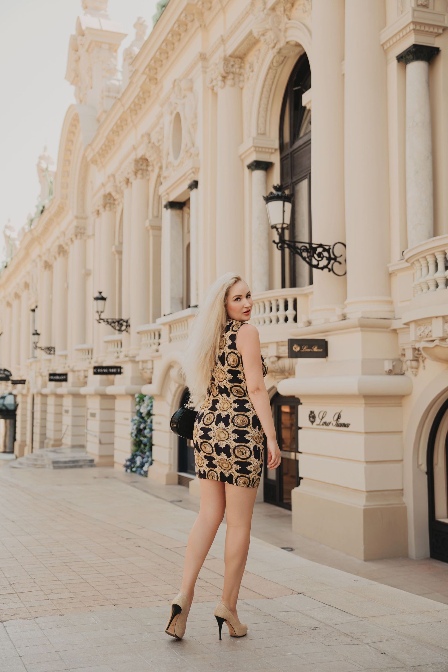 Elegant blonde woman in Versace-print mini dress and heels beside ornate European building.
