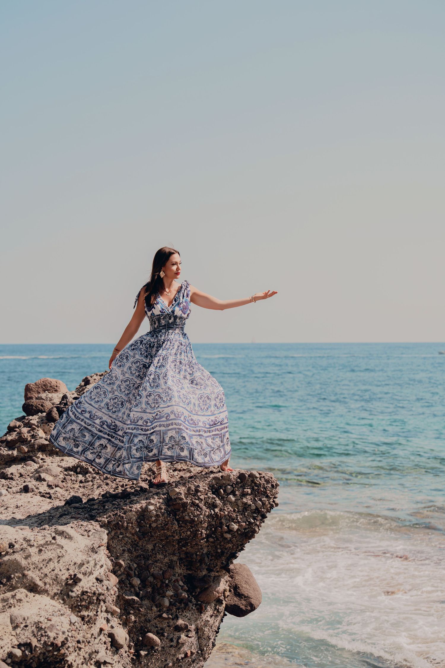 Woman in blue patterned maxi dress standing on rocky seaside cliff with turquoise ocean backdrop.