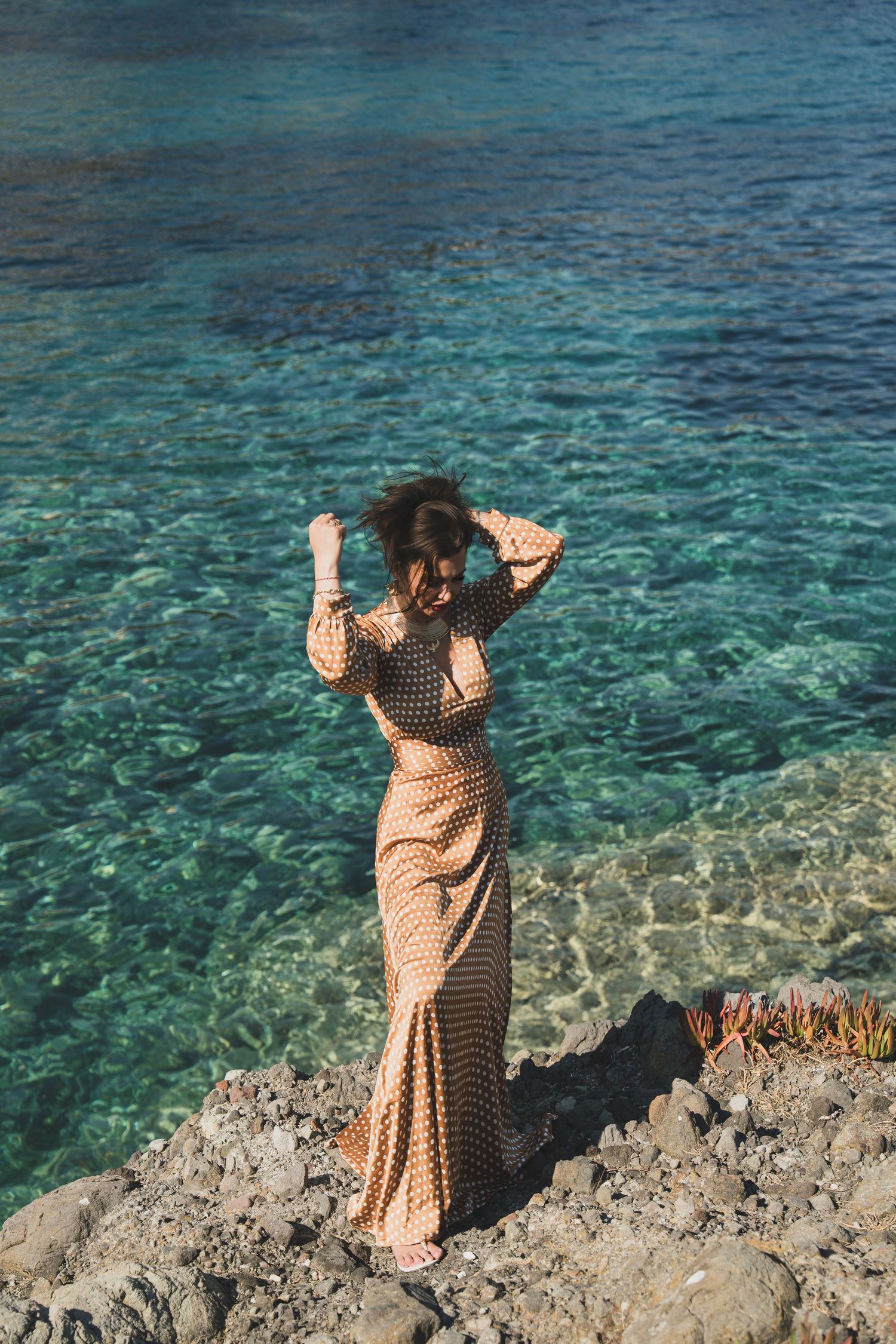 Woman in beige patterned maxi dress with hands in hair on rocky coastal shoreline.