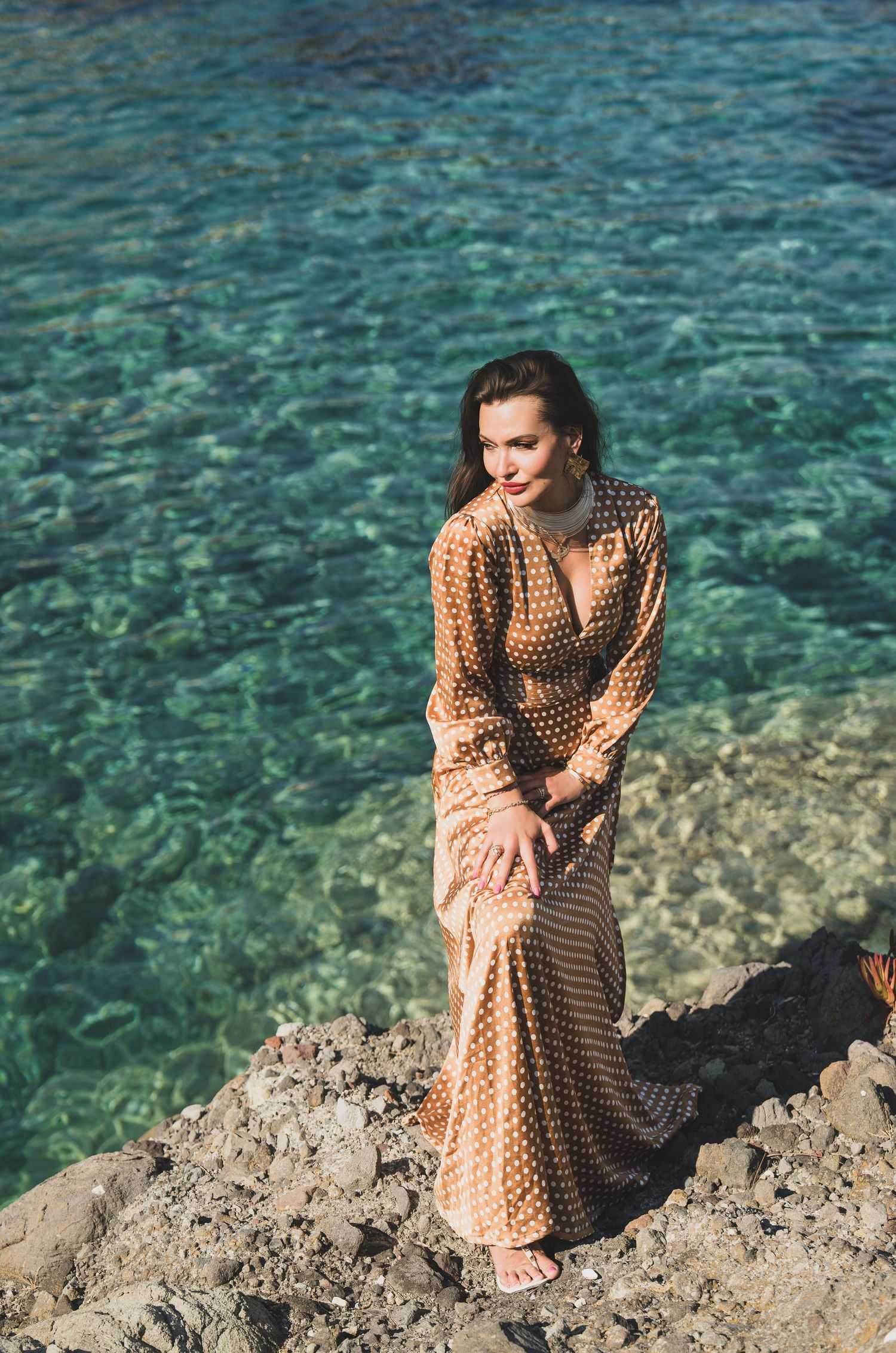 Woman in gold polka dot maxi dress posing on rocky shore with crystal clear turquoise water behind her.