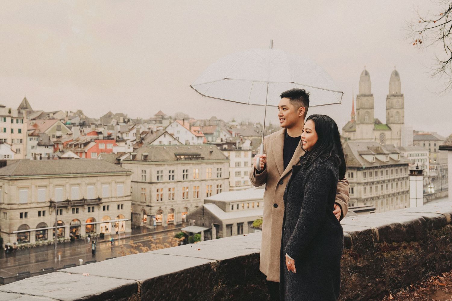 Romantic couple embracing under a transparent umbrella with Zurich cityscape in the background.