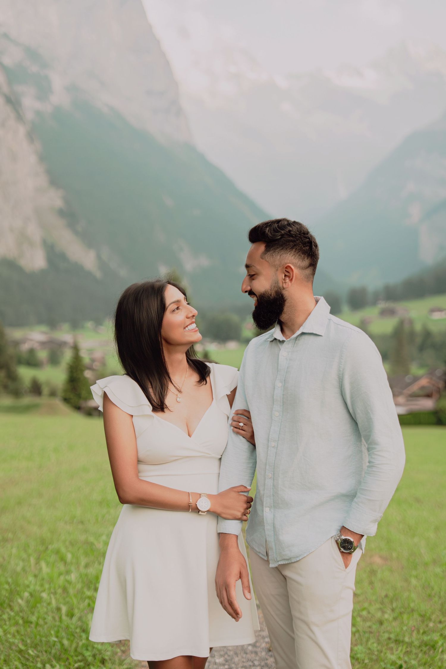 Smiling engaged couple gazing at each other in a lush Swiss alpine meadow with mountain views.