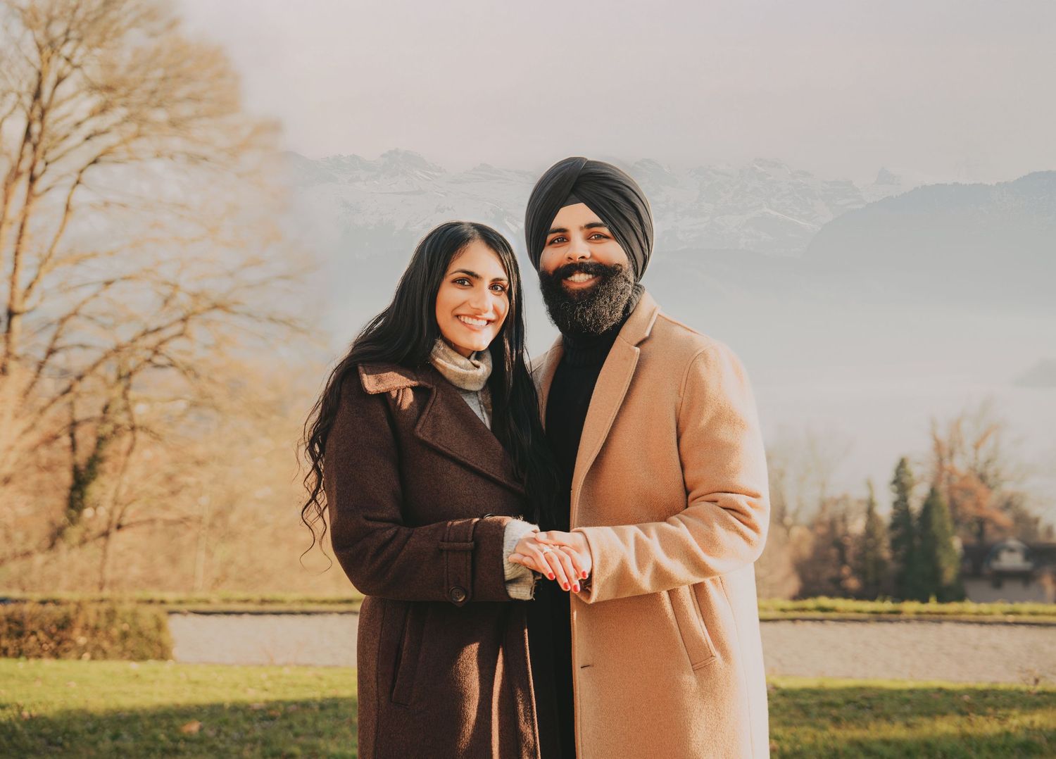 Smiling South Asian couple in winter coats holding hands outdoors with misty mountains in background.