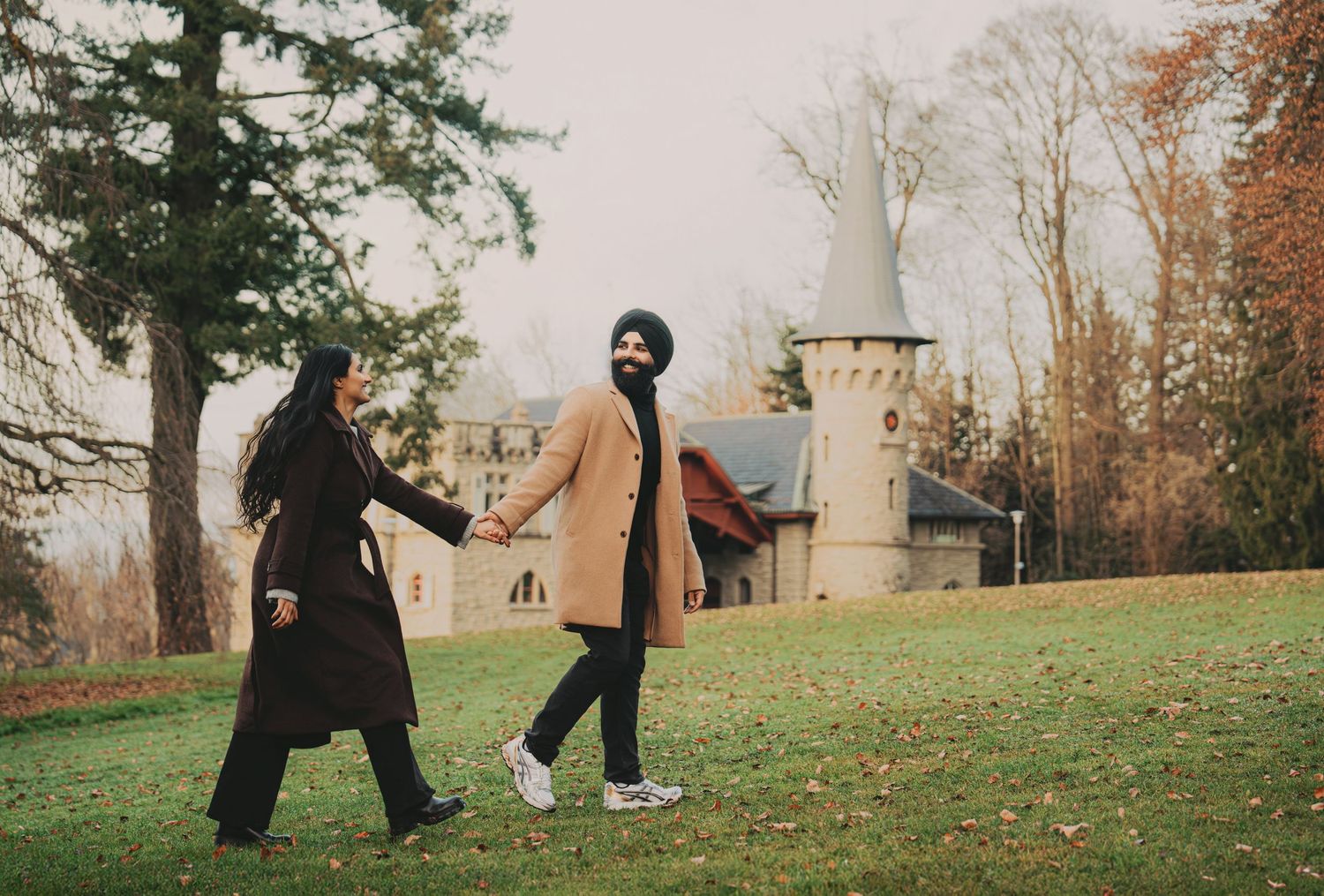 Couple walking hand in hand across a grassy field with a charming castle tower in the background.