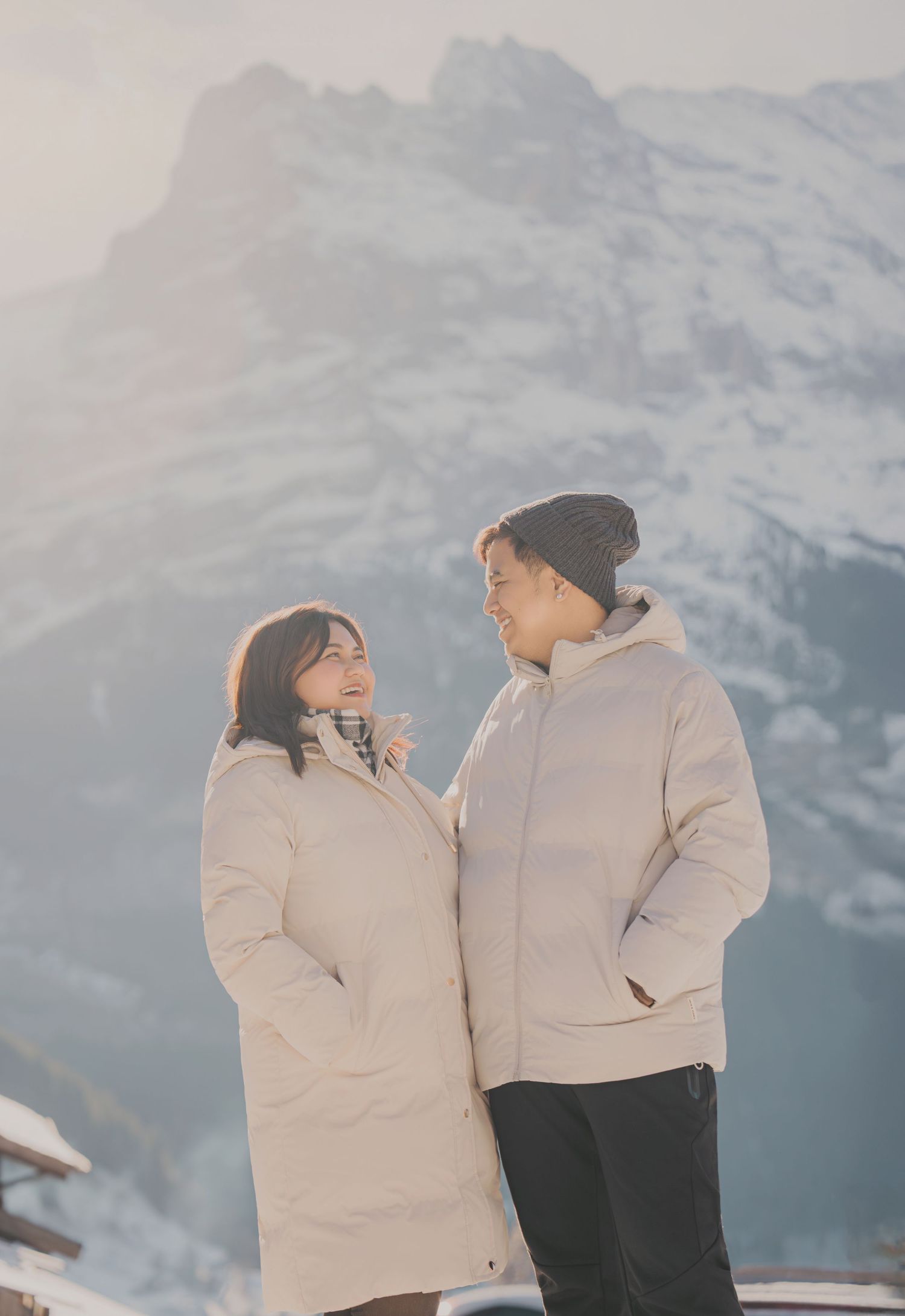 Couple in white winter coats smiling at each other with golden sunlit snowy mountain behind.
