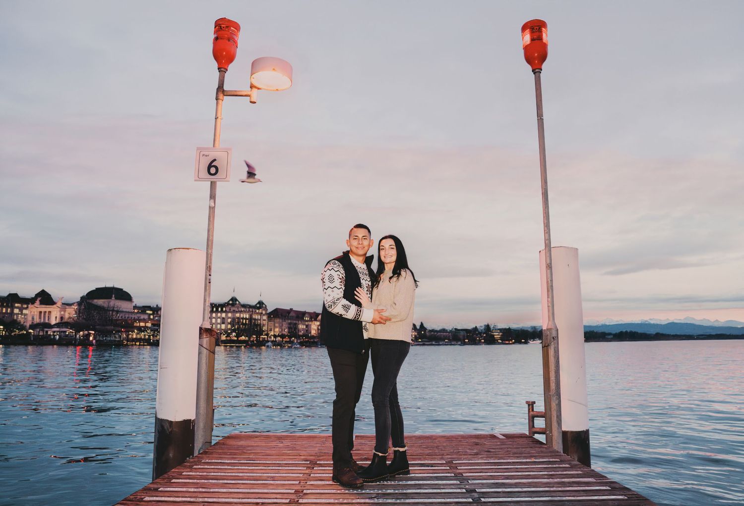 Smiling couple posing together on a lakeside dock with red lamp posts at twilight.