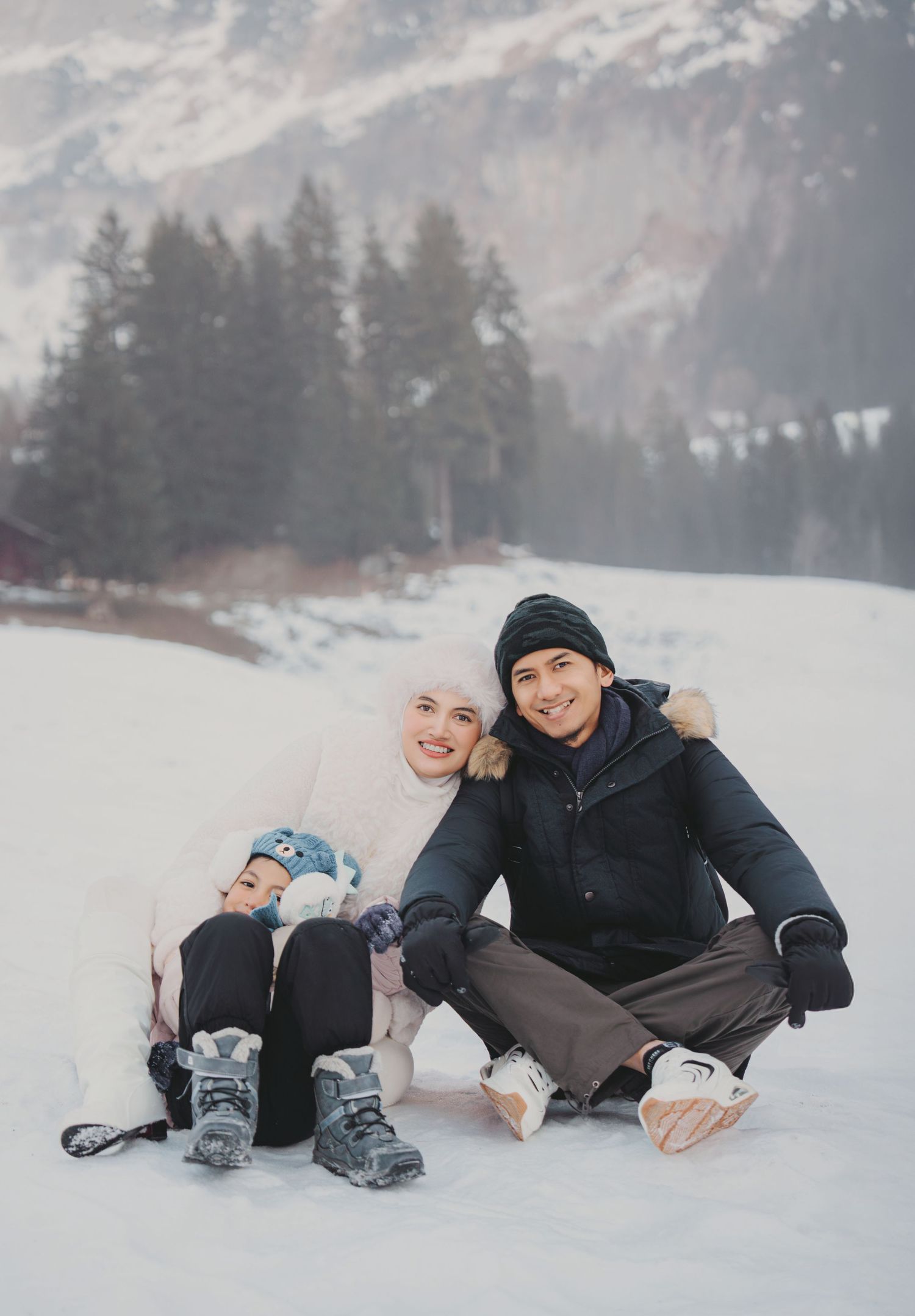 Family of three sitting on snowy ground with mountain and pine trees behind them in winter.