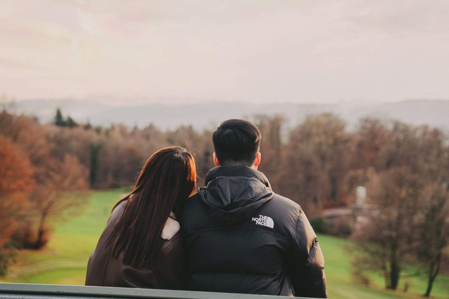 Couple seen from behind leaning on railing overlooking autumn landscape and mountains.
