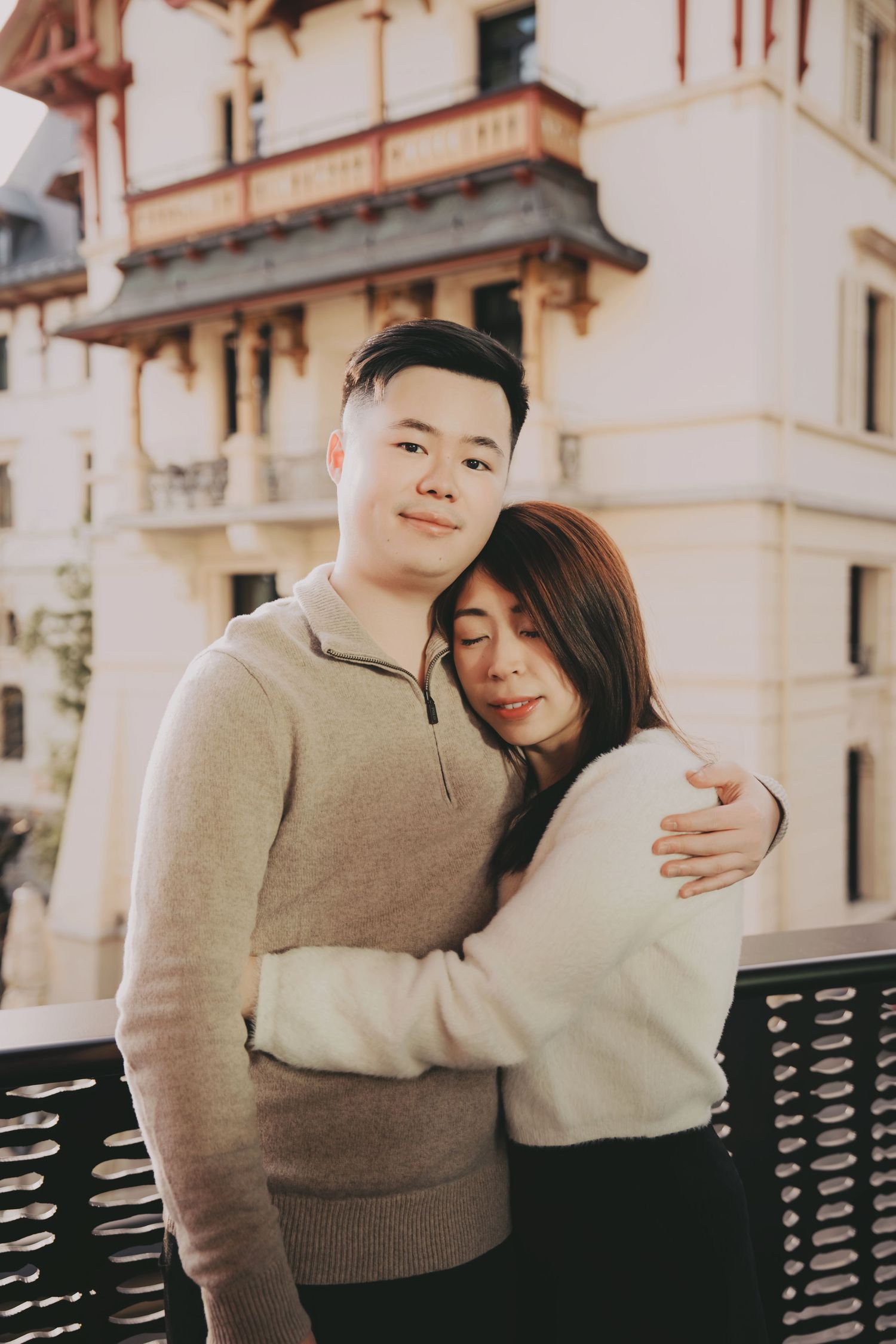 Young Asian couple embracing warmly on a balcony with a classic European building in the soft background.