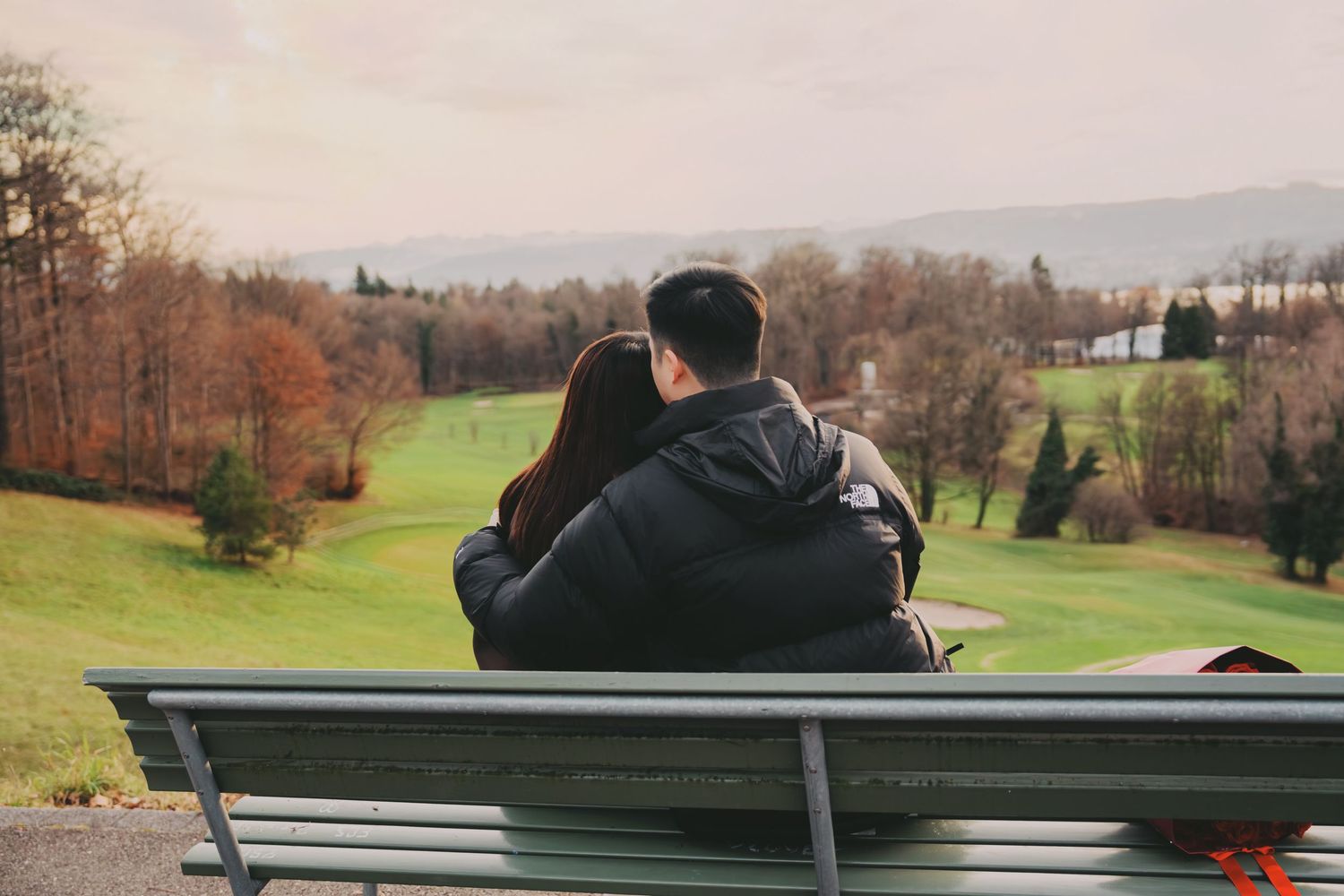 Romantic couple sitting embraced on bench overlooking scenic green golf course and autumn trees.