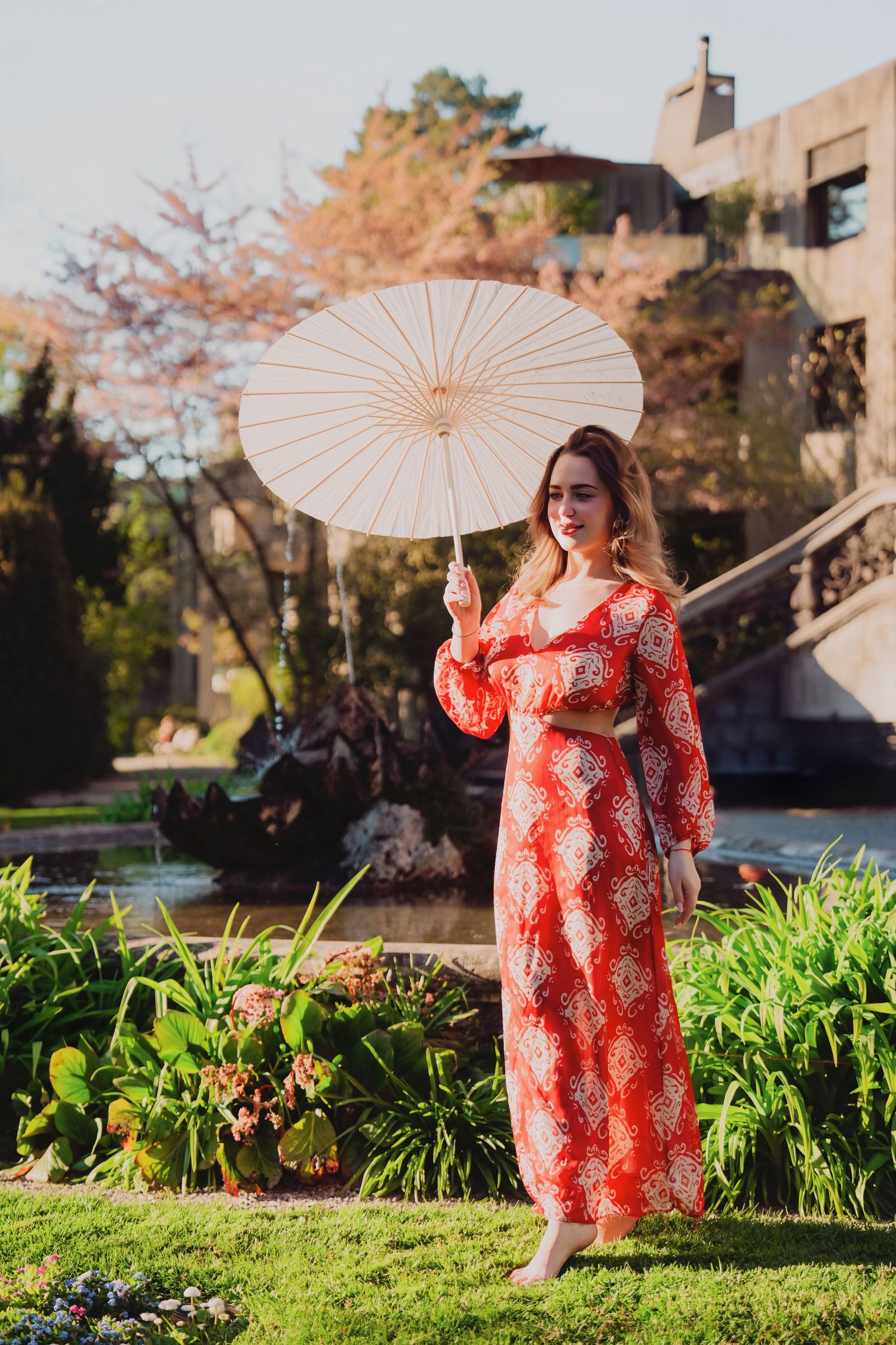 Barefoot woman in red patterned maxi dress stands in garden holding white paper parasol umbrella.