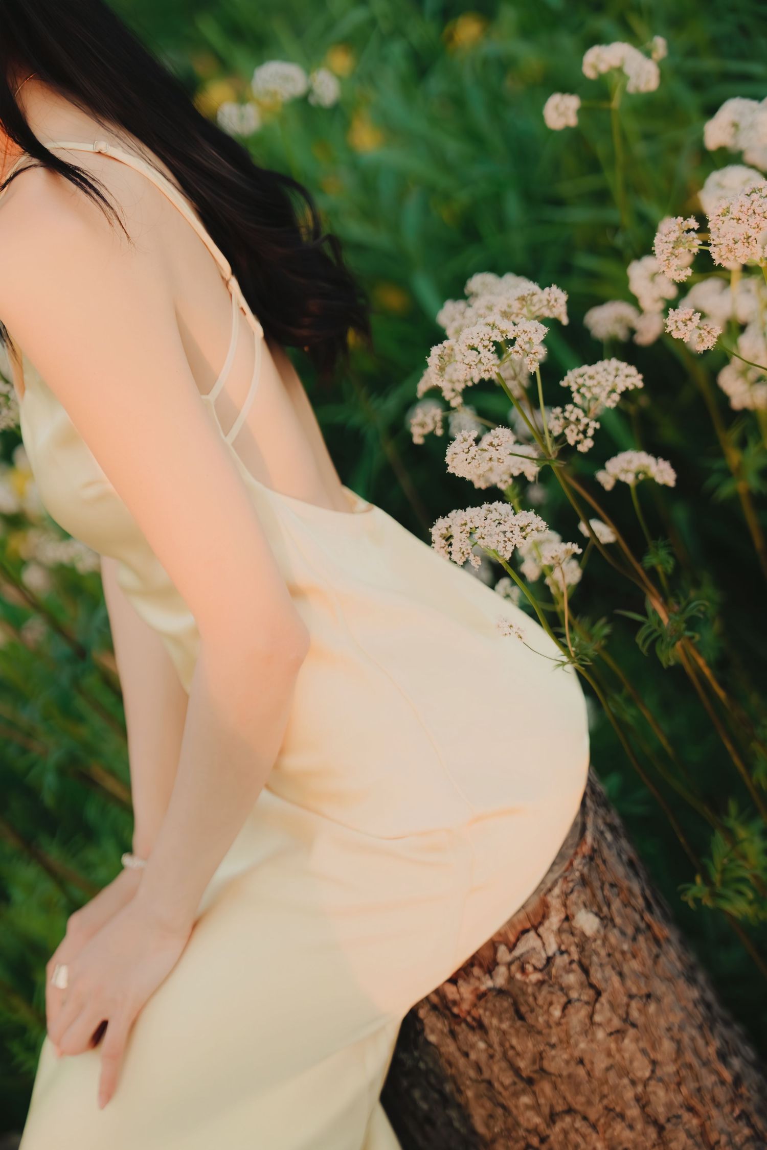 Woman in elegant pale yellow slip dress sitting on a tree stump surrounded by white wildflowers.