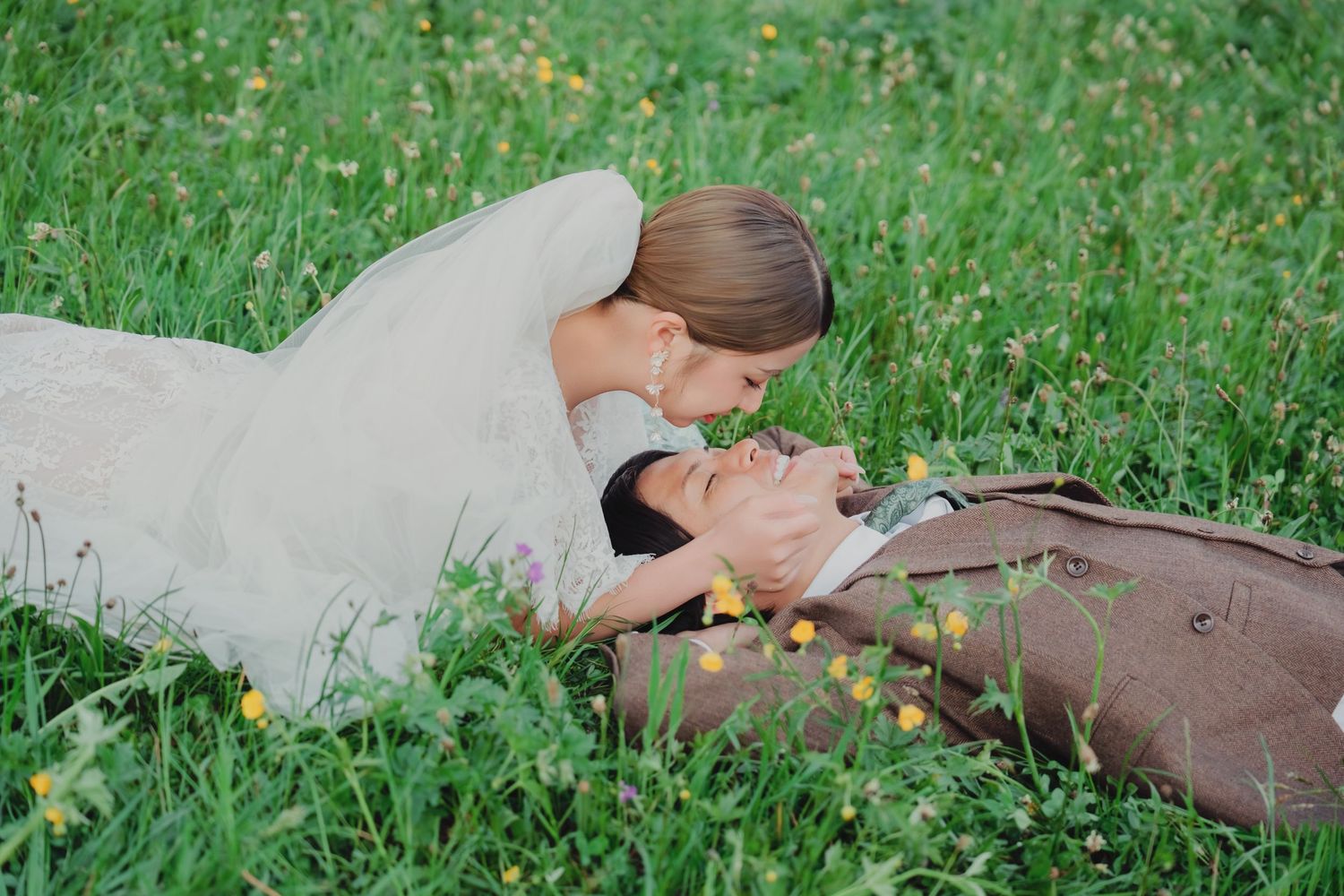 Bride tenderly leaning over smiling groom lying in lush green Swiss alpine meadow.