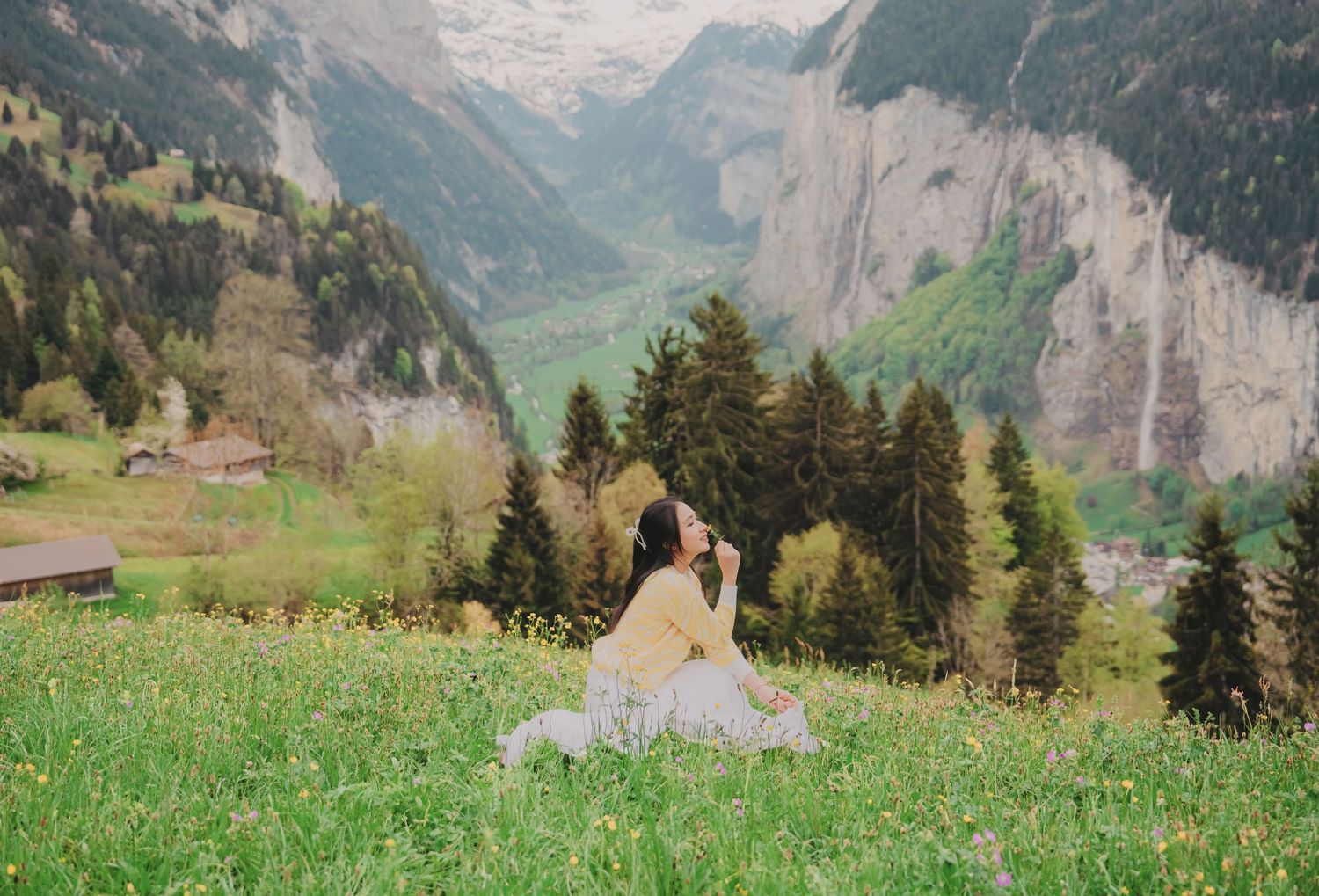 Woman in yellow outfit sitting in green alpine meadow with Swiss mountain valley and waterfall view.