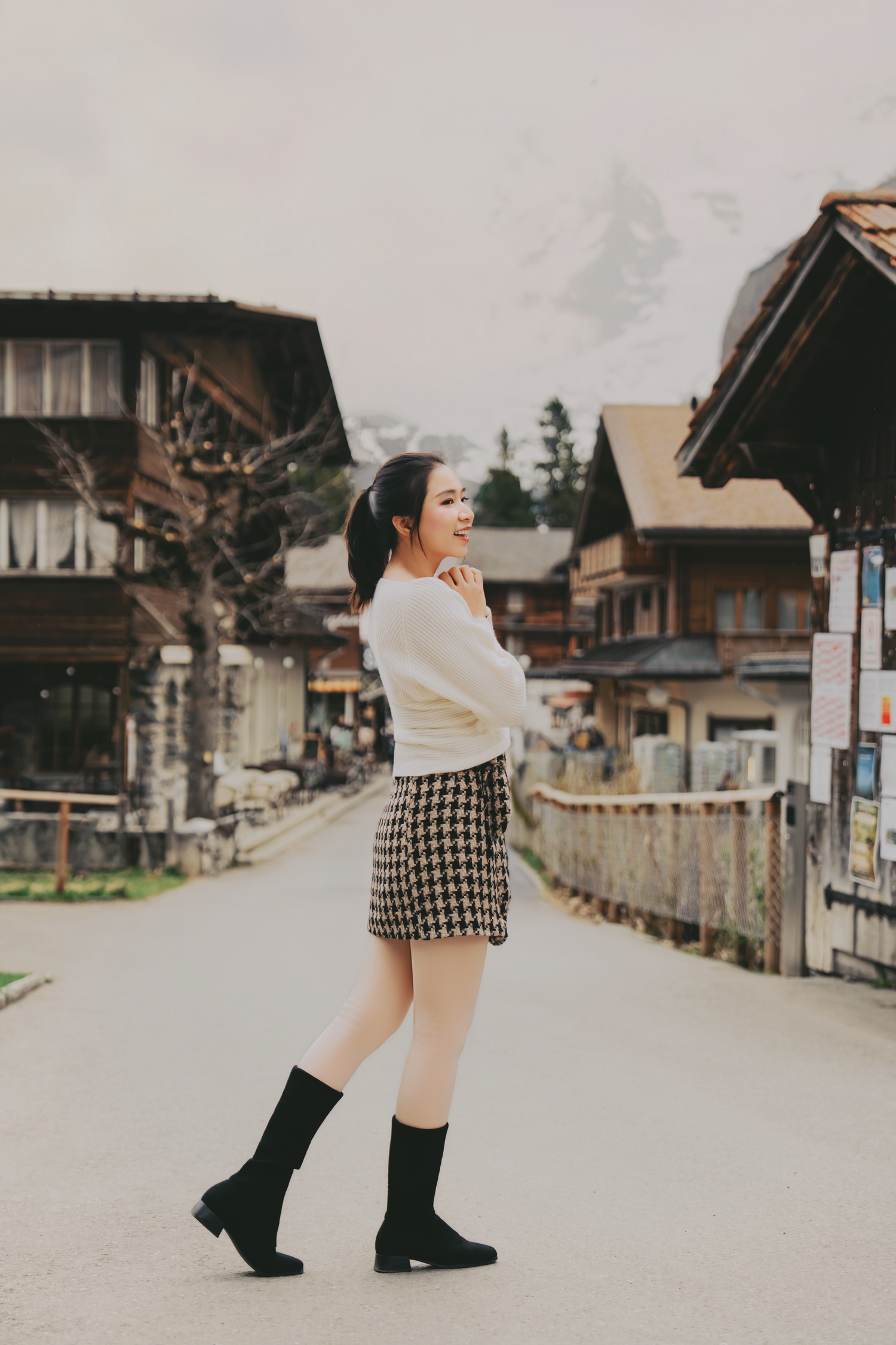 Stylish Asian woman in white sweater and houndstooth skirt posing on charming Swiss village street.