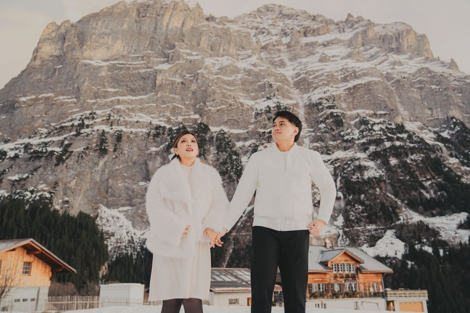 Couple holding hands in white winter outfits with snow-covered Dolomites mountain backdrop.