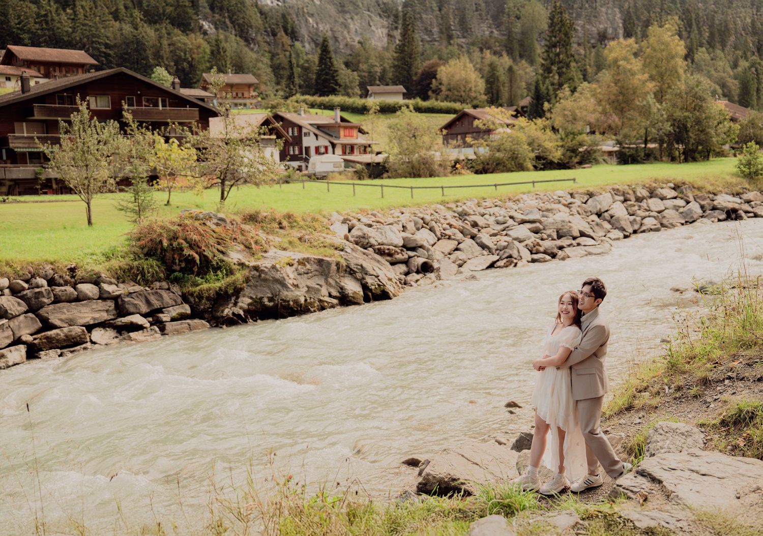 Couple embracing by a turquoise river with traditional Swiss alpine village scenery.