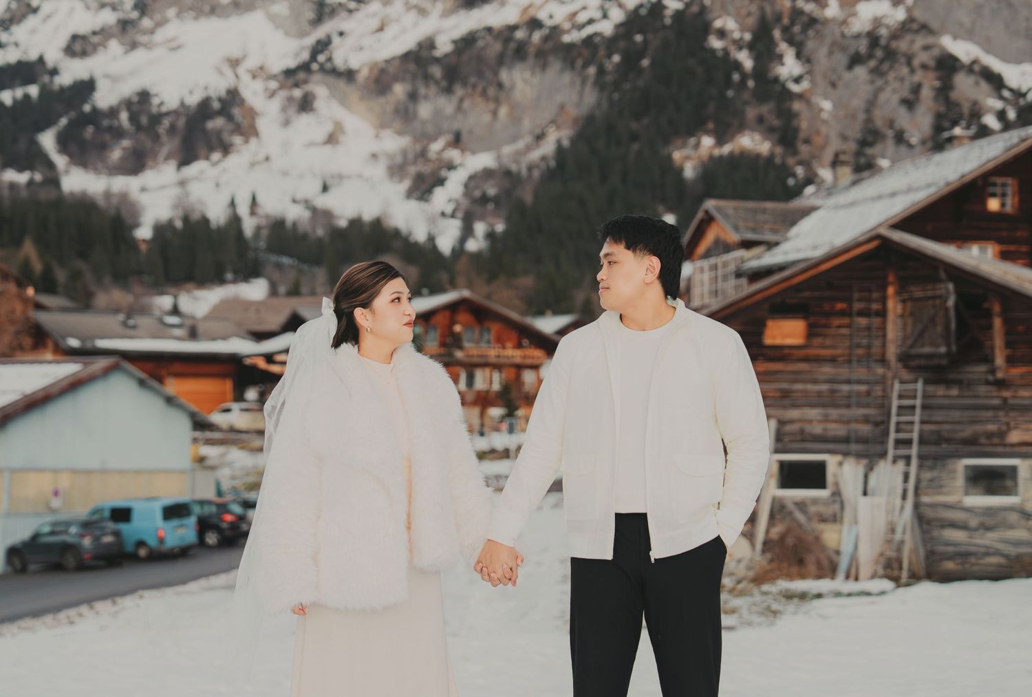 Couple in white winter outfits holding hands and smiling in a snowy Swiss mountain village setting.