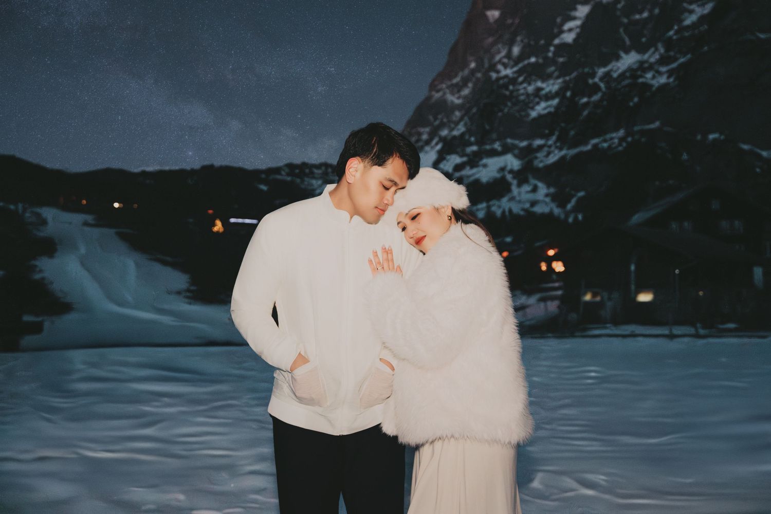 Romantic couple embracing in white outfits against snowy mountain backdrop at night.