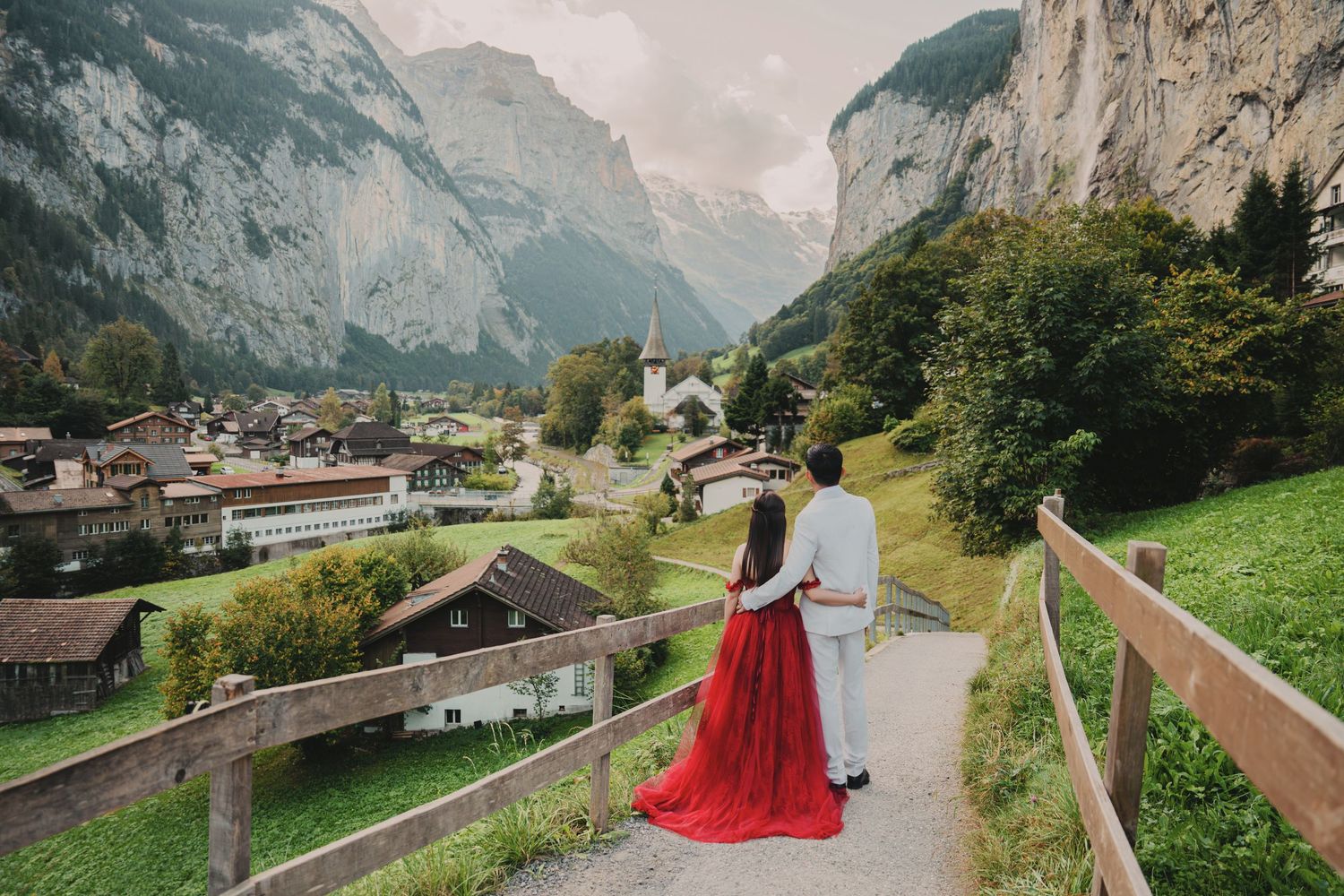 Couple in red dress and white suit walking on a path with stunning Swiss village and mountain view.